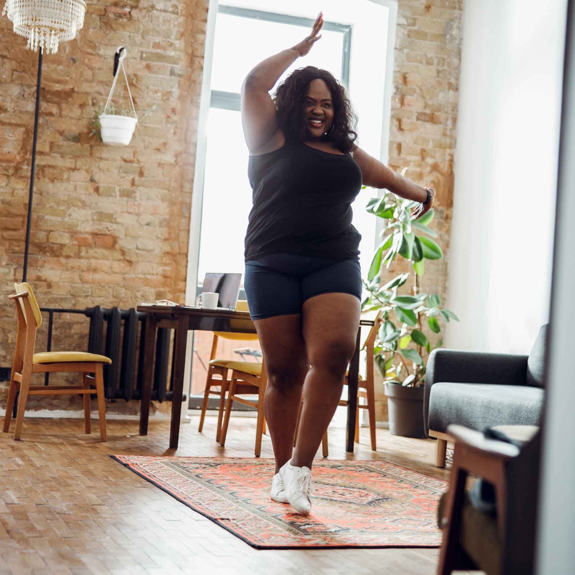 A person joyfully dancing in a cozy living room with brick walls, a plant, and wooden furniture.
