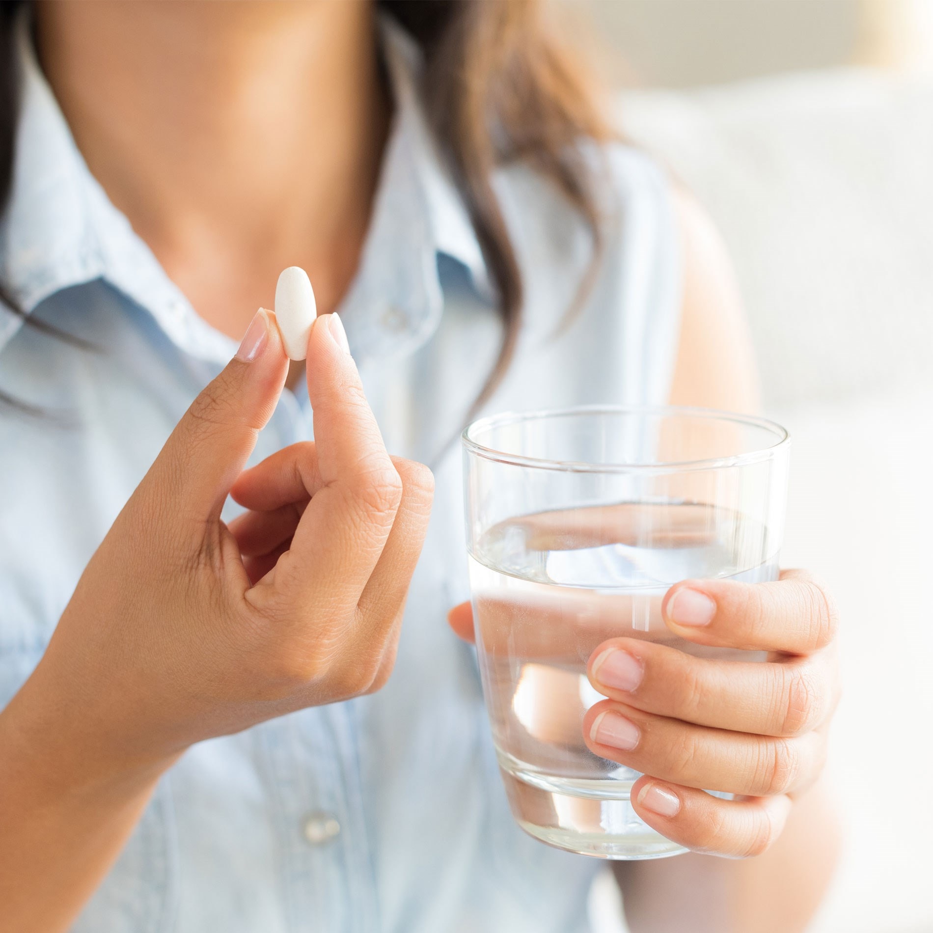 Person holding a white pill in one hand and a glass of water in the other, wearing a sleeveless denim shirt.
