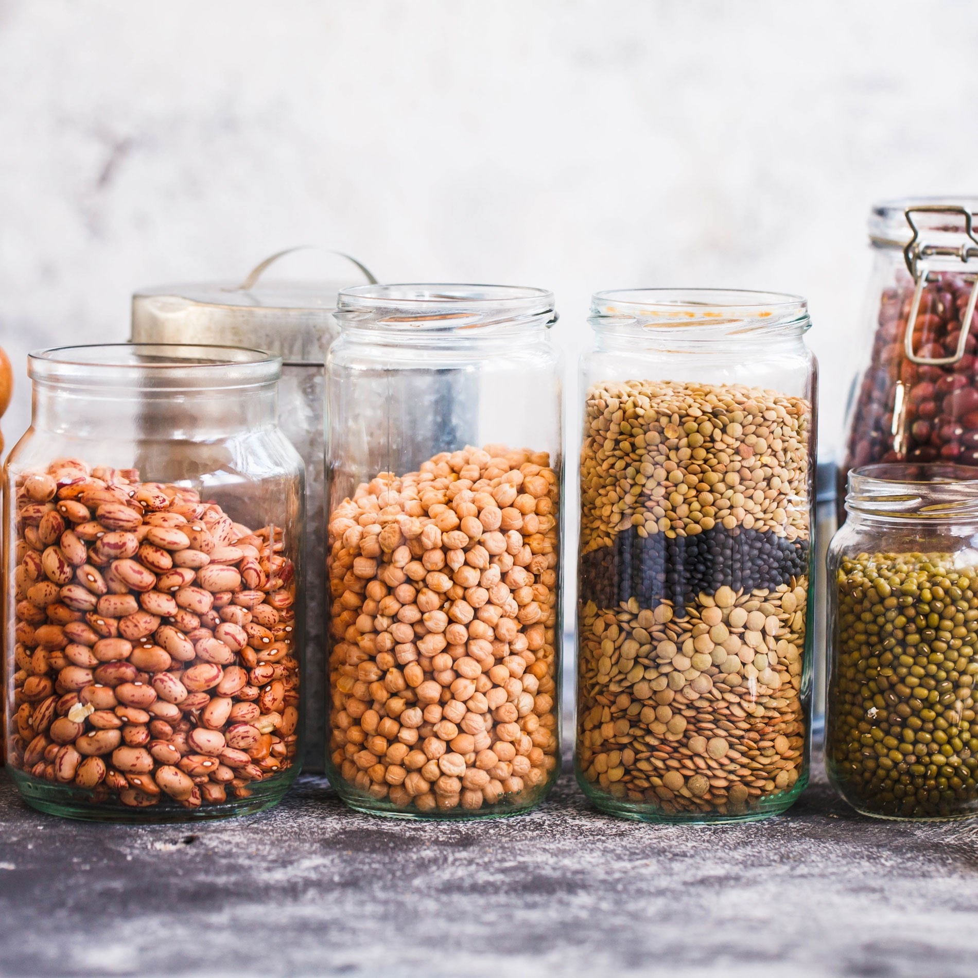 Glass jars filled with various dried legumes, including beans, chickpeas, lentils, and peas, arranged on a textured surface.