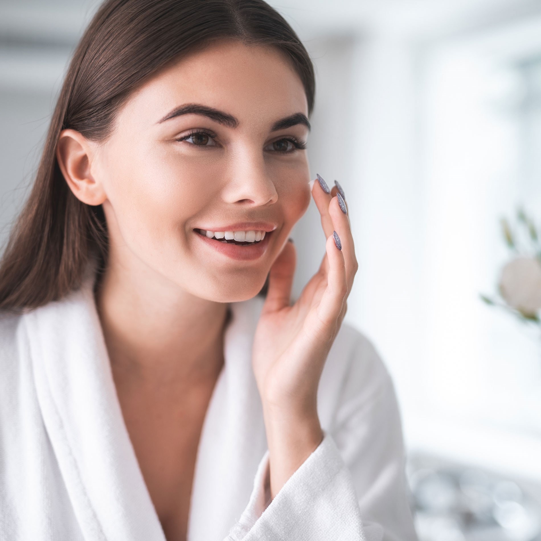 Woman in a white robe smiling and applying skincare product to her cheek in a bright room.