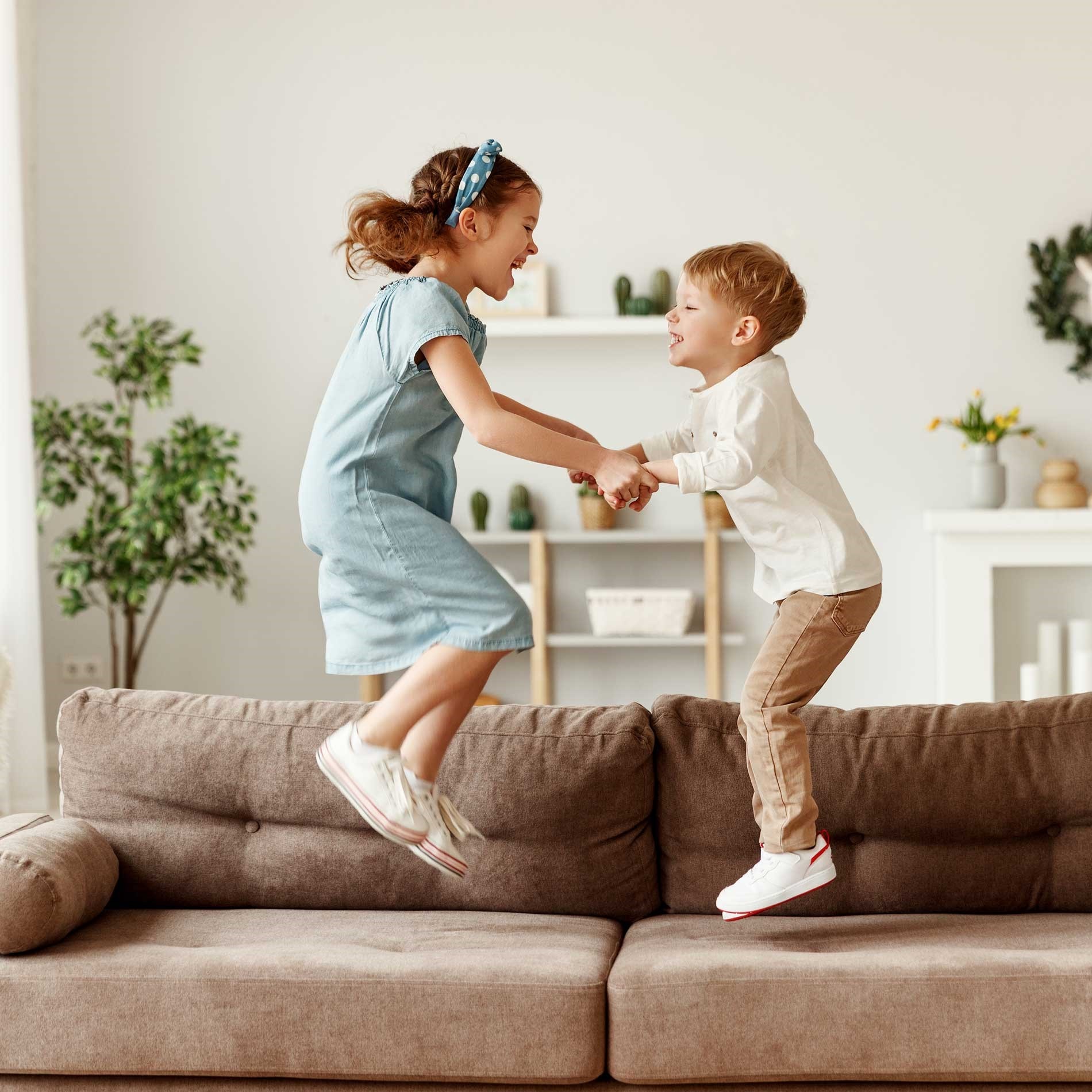 Two children joyfully jumping on a brown couch, holding hands. The room has a light decor with plants and shelves in the background.