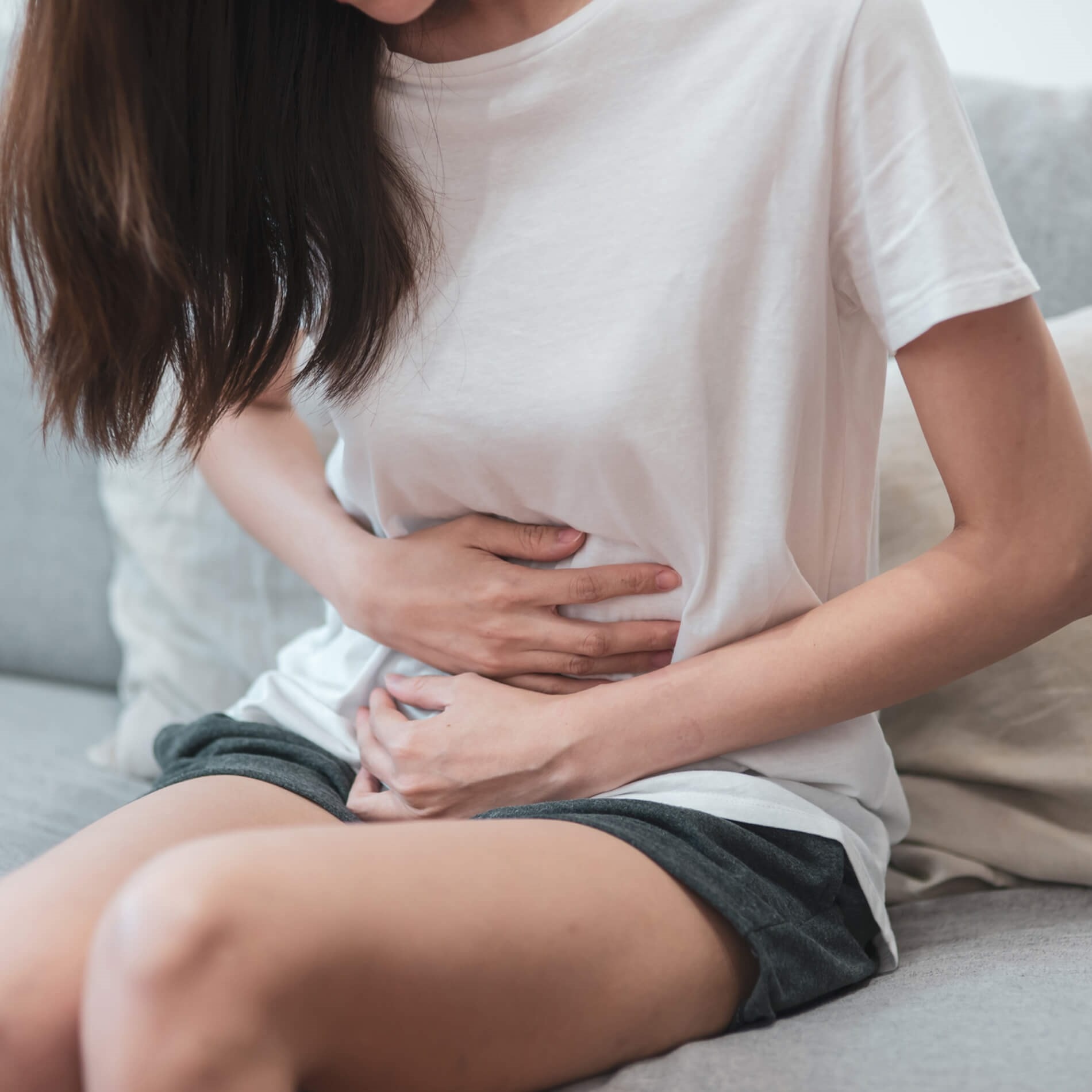 A woman sitting on a couch, holding her stomach with both hands, appearing to be in discomfort.