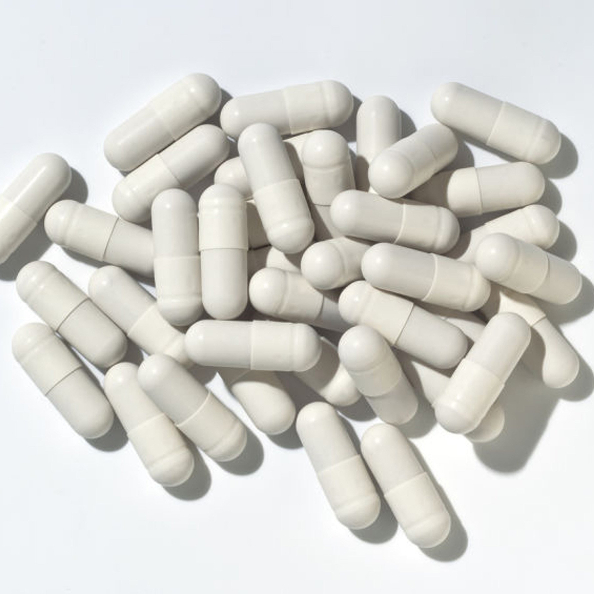 A pile of white capsules scattered on a white surface, with shadows visible, suggesting a bright light source.