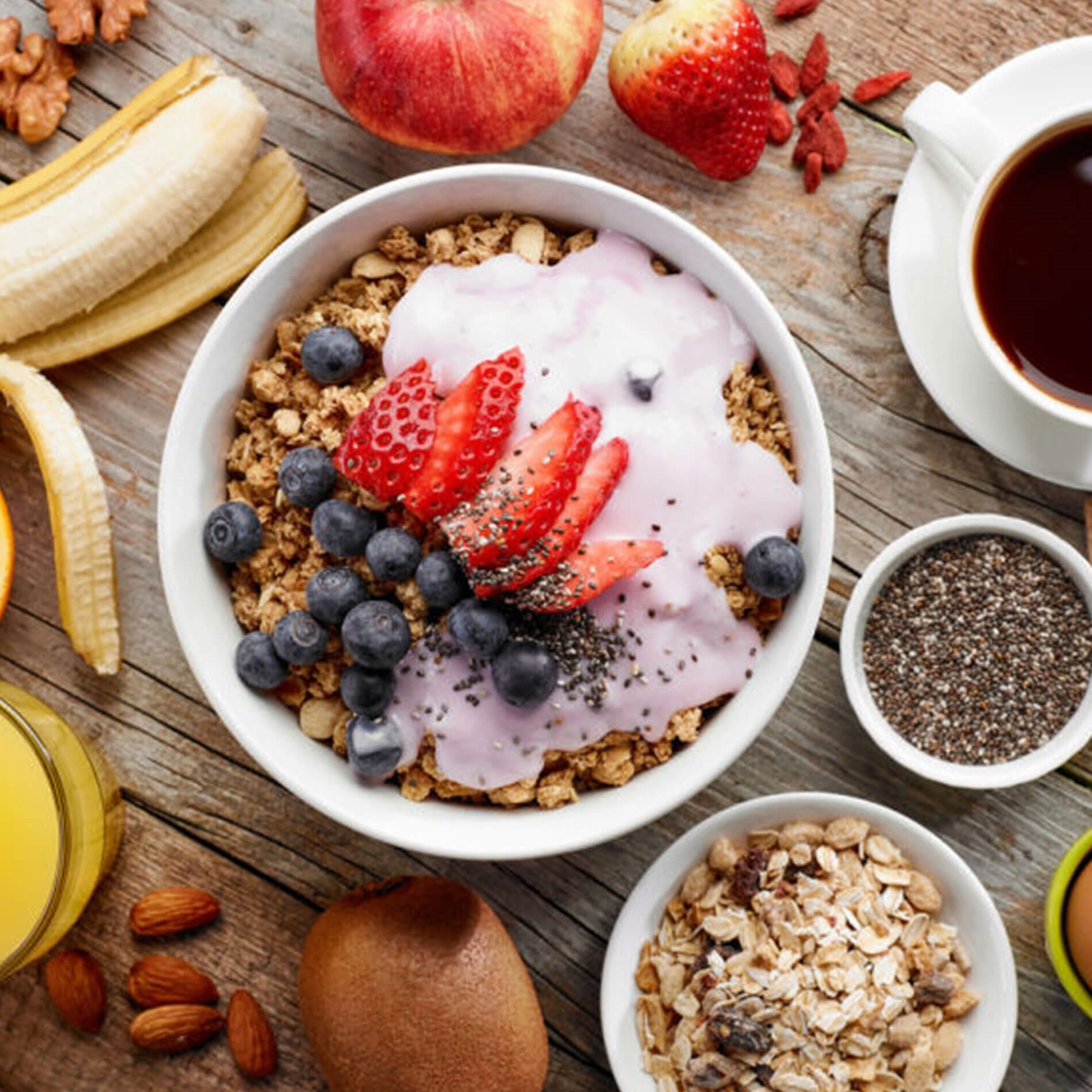 A bowl of granola with yogurt, strawberries, blueberries, and chia seeds, surrounded by fruit, nuts, orange juice, and coffee on a wooden table.