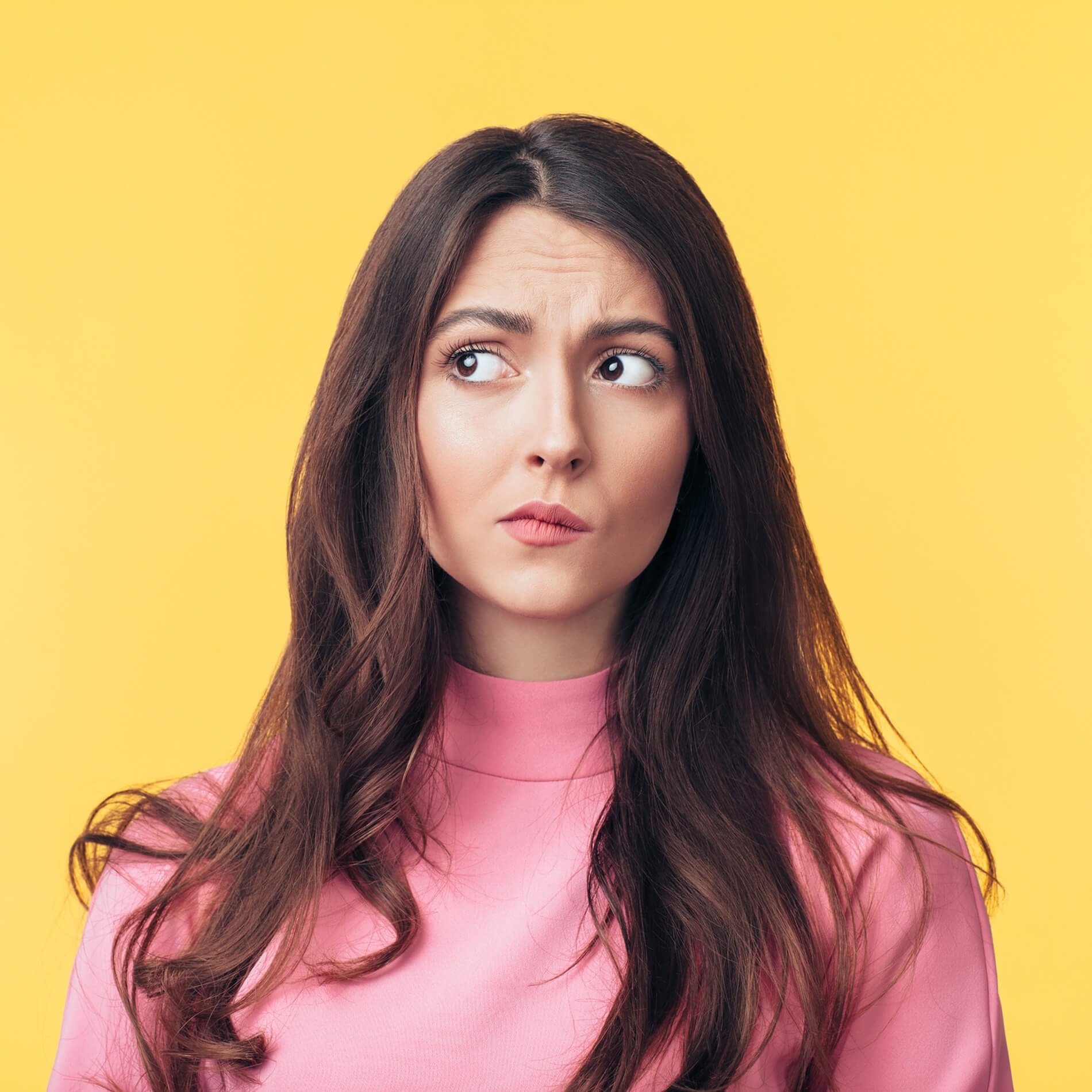 Woman with long brown hair in a pink top looks to the side with a pensive expression against a yellow background.