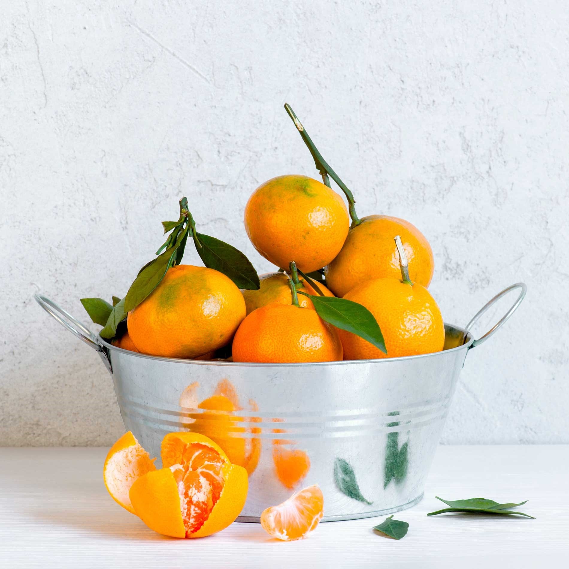 A metal bucket filled with ripe oranges, some with leaves, on a white surface. Peeled orange segments are placed nearby.