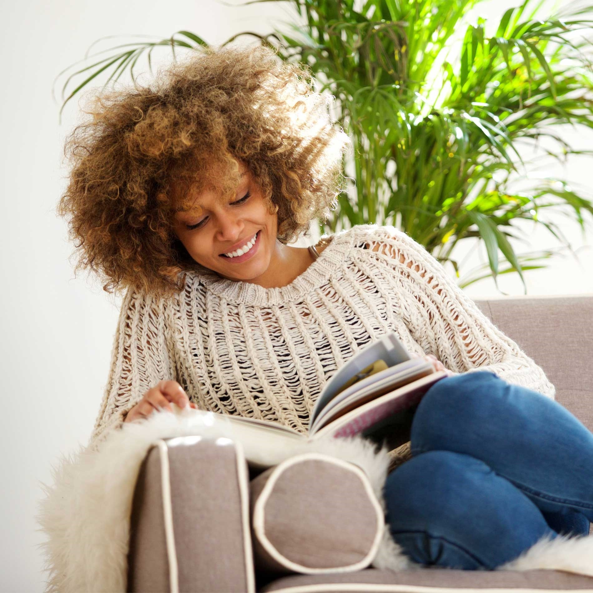 Woman with curly hair, wearing a knit sweater, sitting on a couch reading a book, smiling, with a green plant in the background.