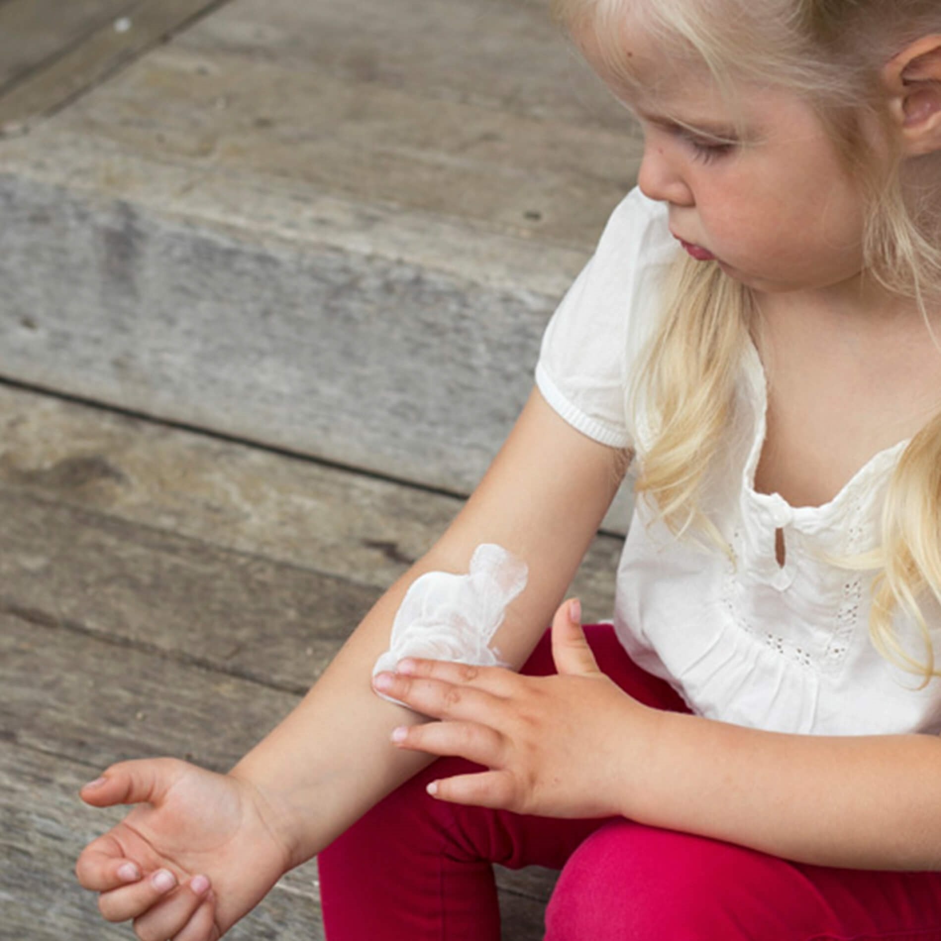 Young girl with blonde hair applies a bandage to her arm while sitting on wooden steps, wearing a white shirt and red pants.