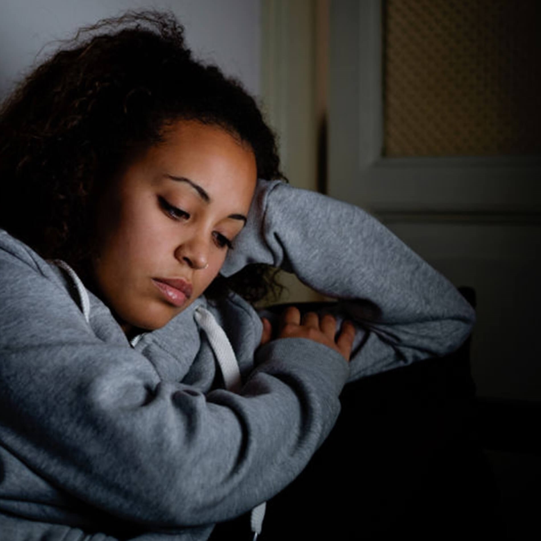 A person in a gray hoodie sits with a thoughtful expression, resting their head on their hand in a dimly lit room.