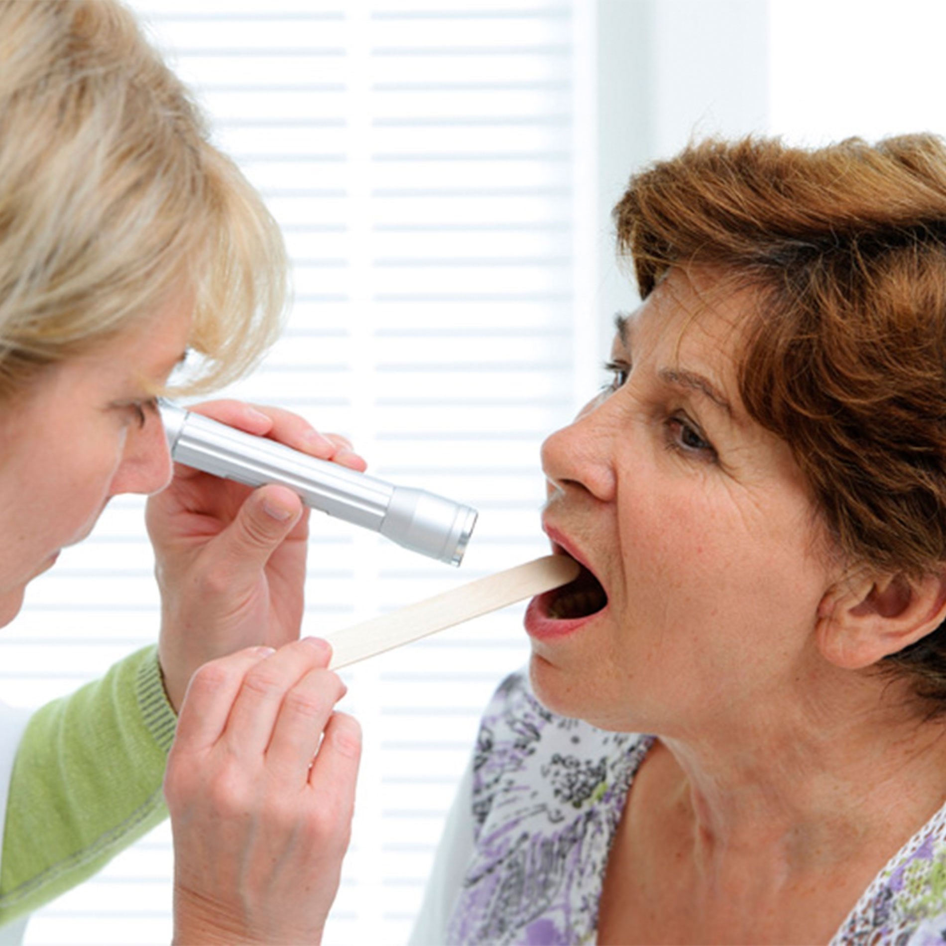 A healthcare professional examines a woman's throat using a flashlight and tongue depressor in a clinical setting.