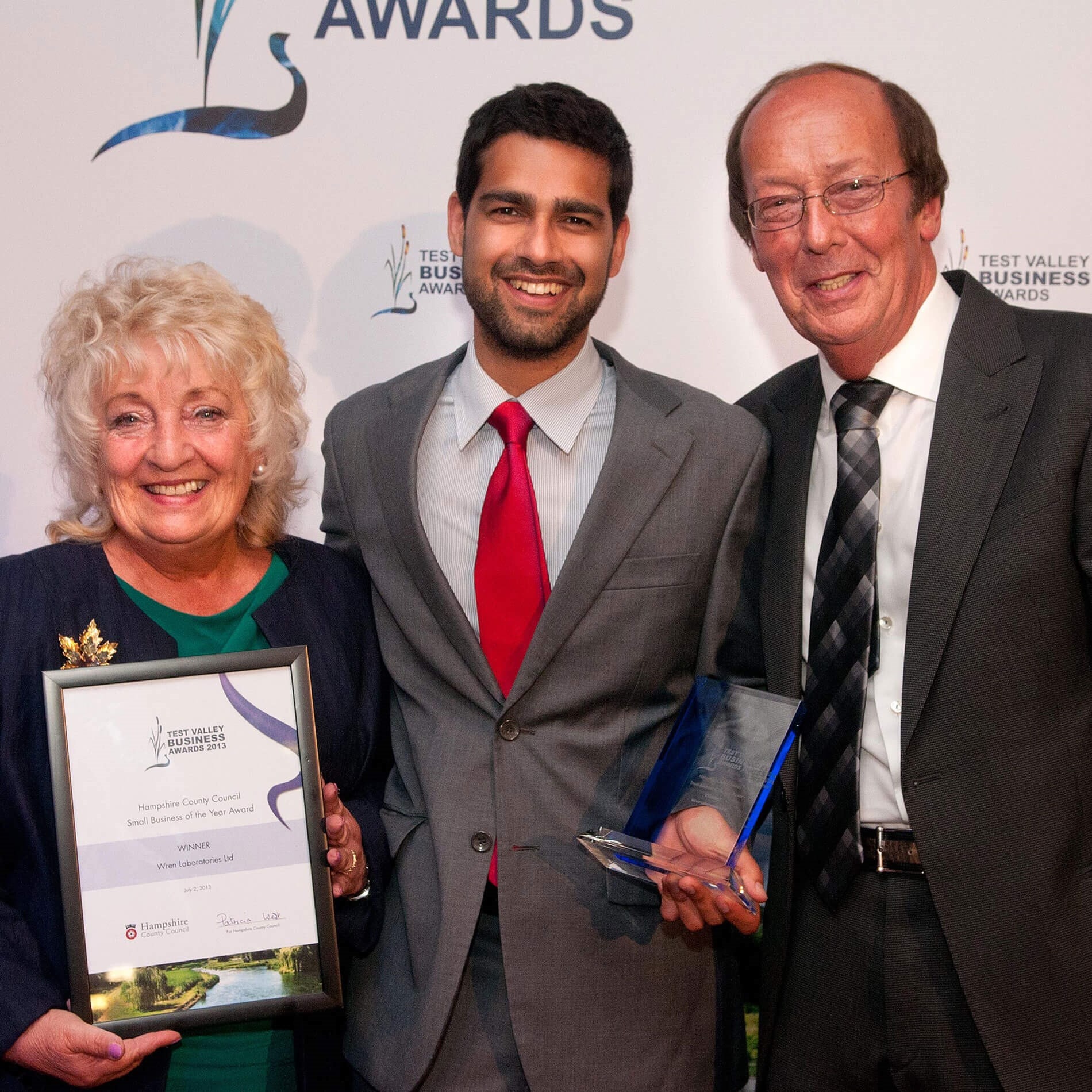 Three people smiling at an awards event; one holds a framed certificate, another holds a glass trophy.