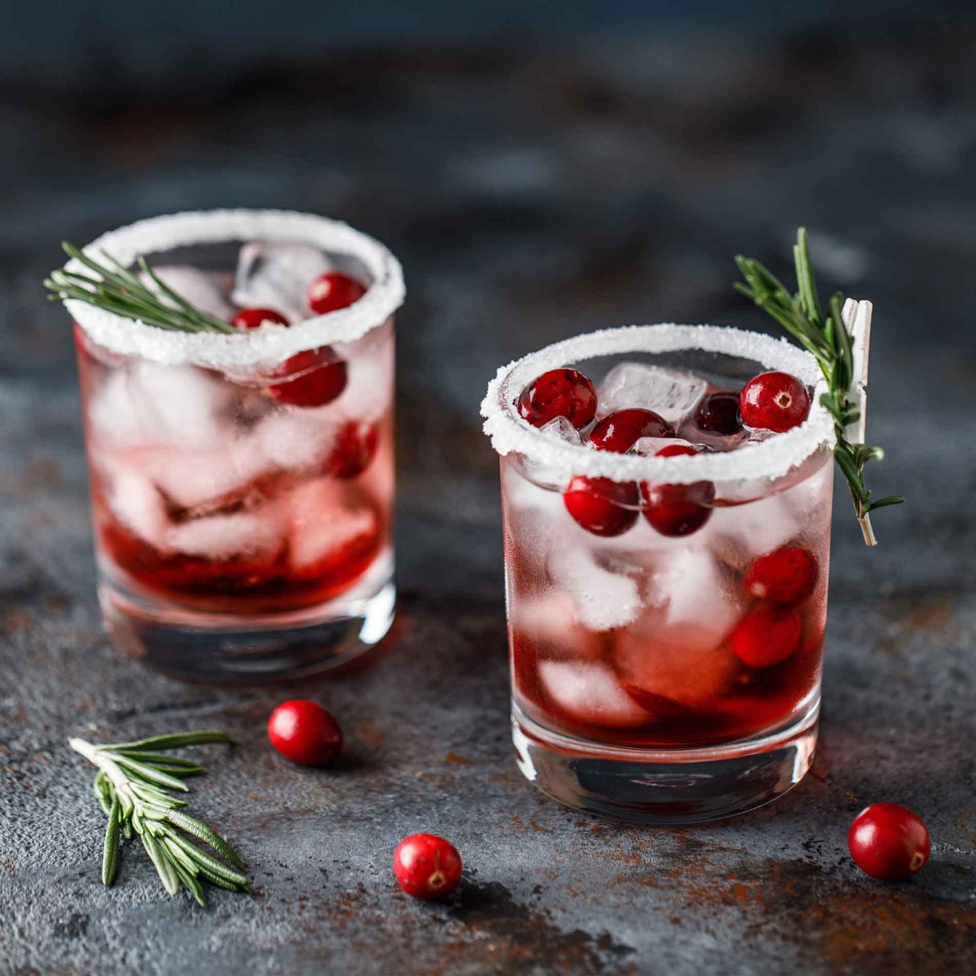 Two glasses of cranberry cocktails with ice, sugar rims, garnished with rosemary sprigs and cranberries, on a dark textured surface.