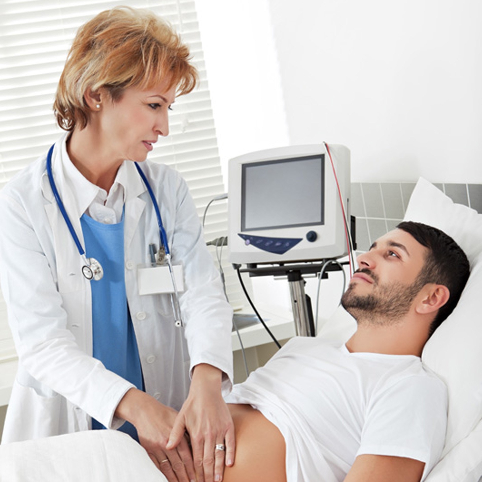 Doctor examining a male patient lying on a hospital bed, with medical equipment in the background.