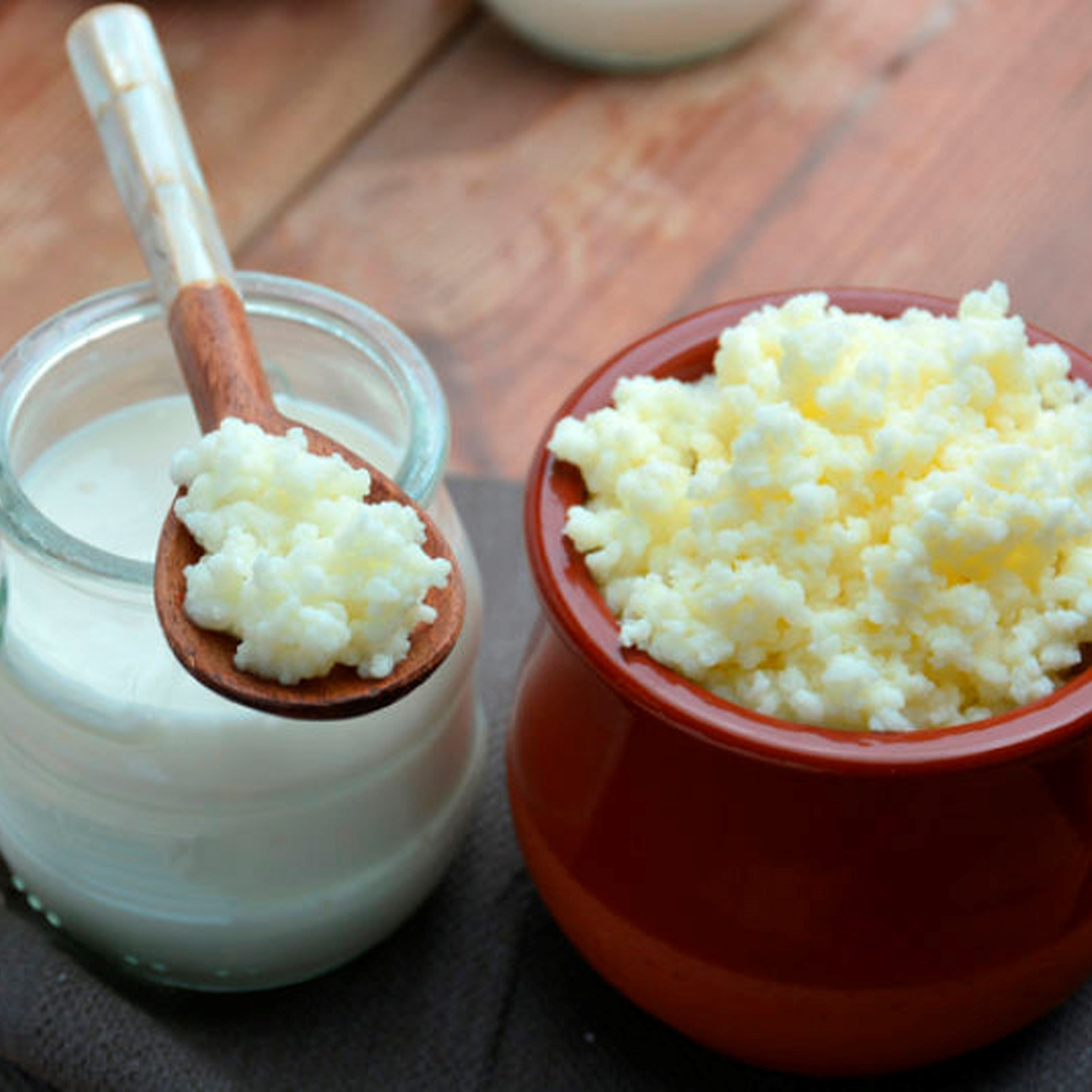 A jar of kefir with a wooden spoon resting on top, and a brown bowl filled with kefir grains, on a wooden surface.