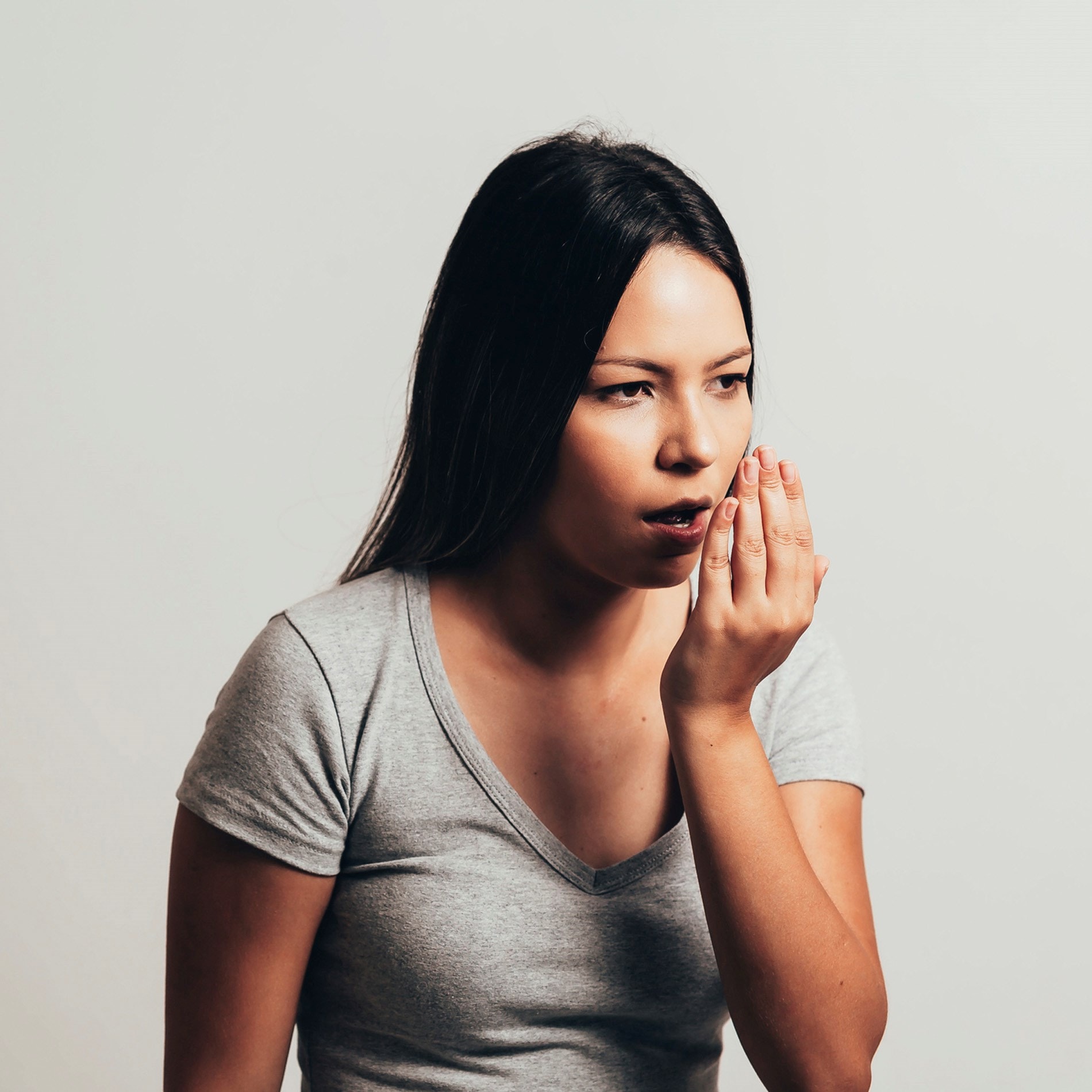 Woman with long hair in a gray shirt checking her breath by covering her mouth with her hand, standing against a plain background.