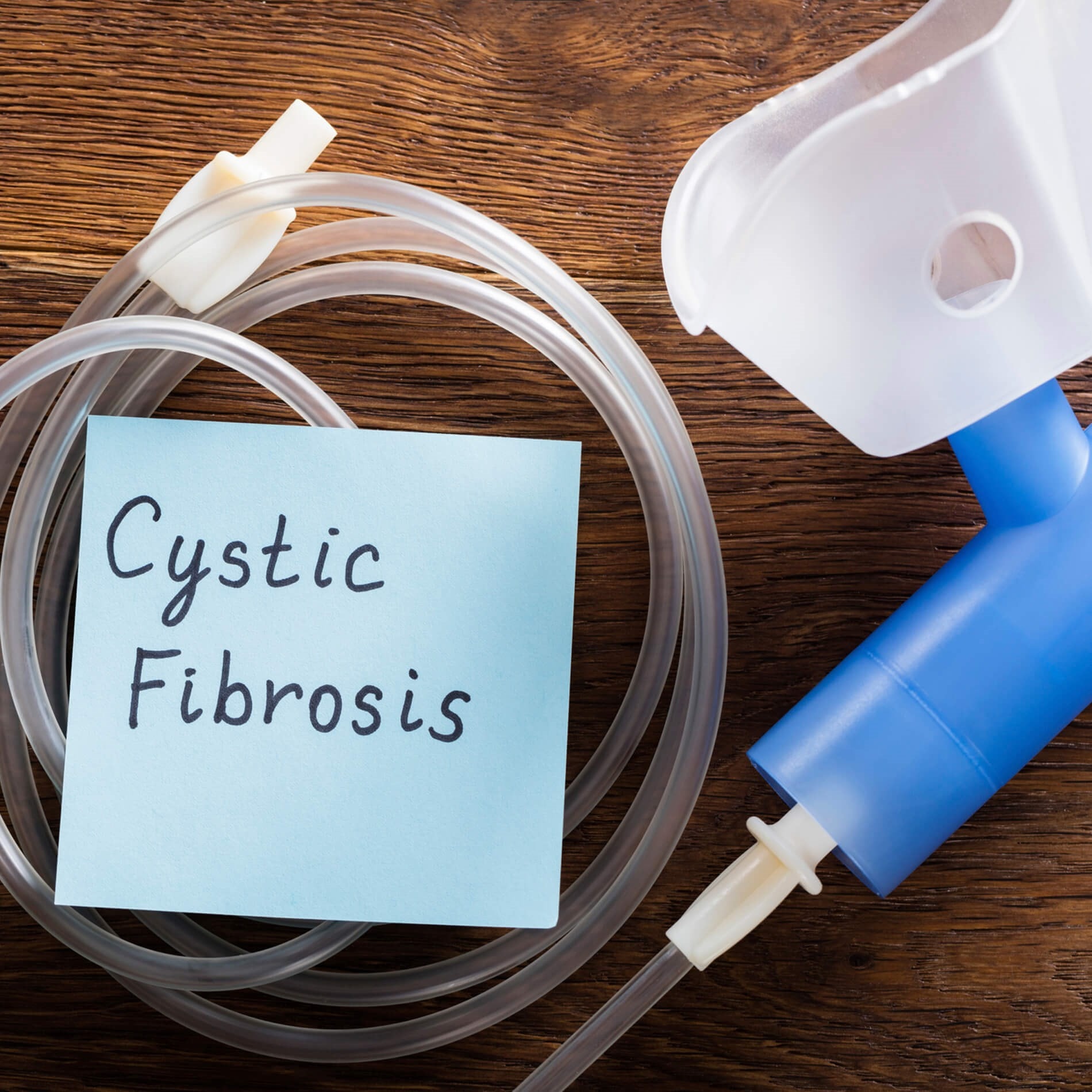 A nebulizer with a blue mouthpiece and clear tubing sits next to a sticky note reading "Cystic Fibrosis" on a wooden surface.