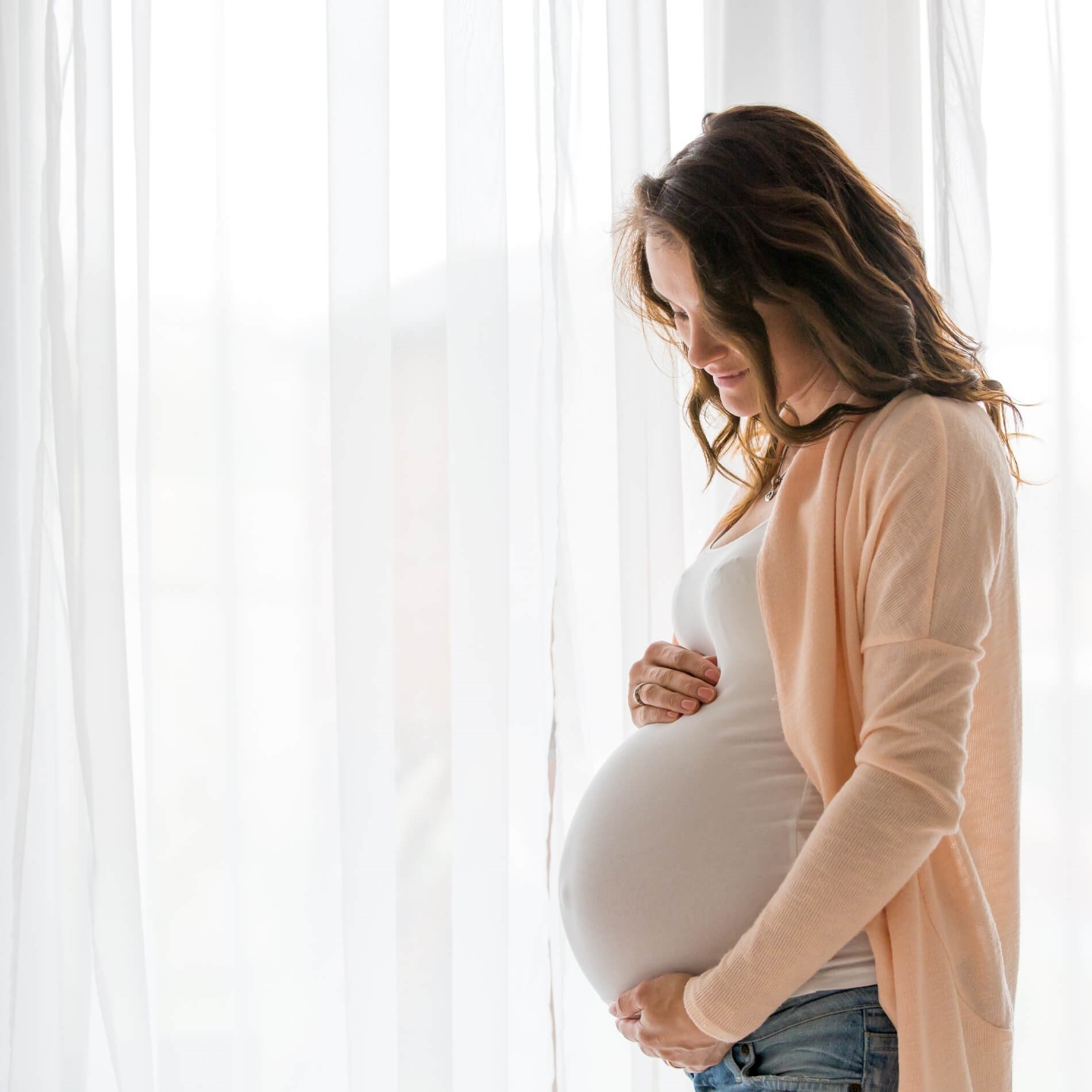 Pregnant woman in a light cardigan cradles her belly, standing by sheer white curtains, looking down with a gentle expression.