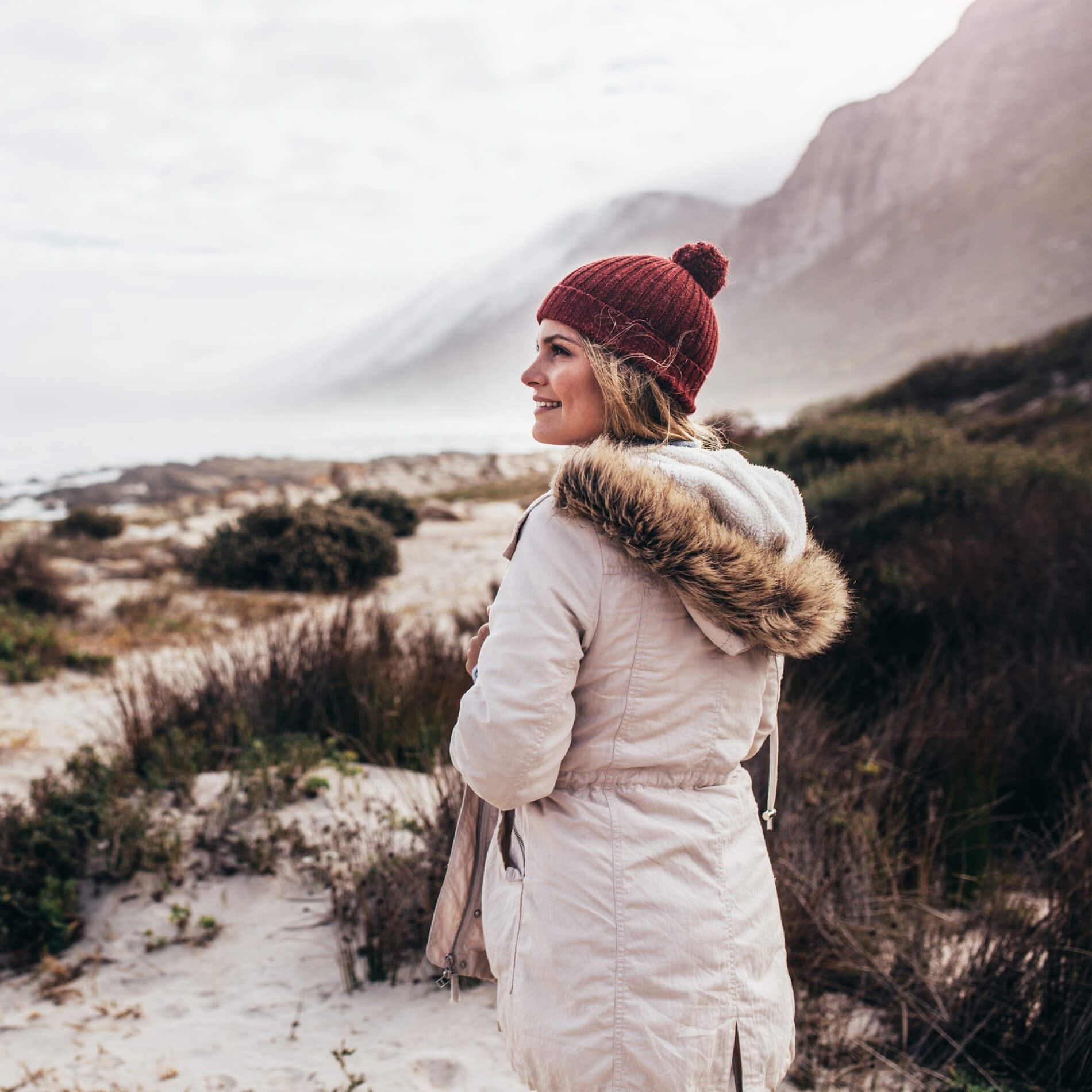 Woman in a fur-lined coat and red beanie stands on a sandy, shrub-covered path by the sea, with misty mountains in the background.