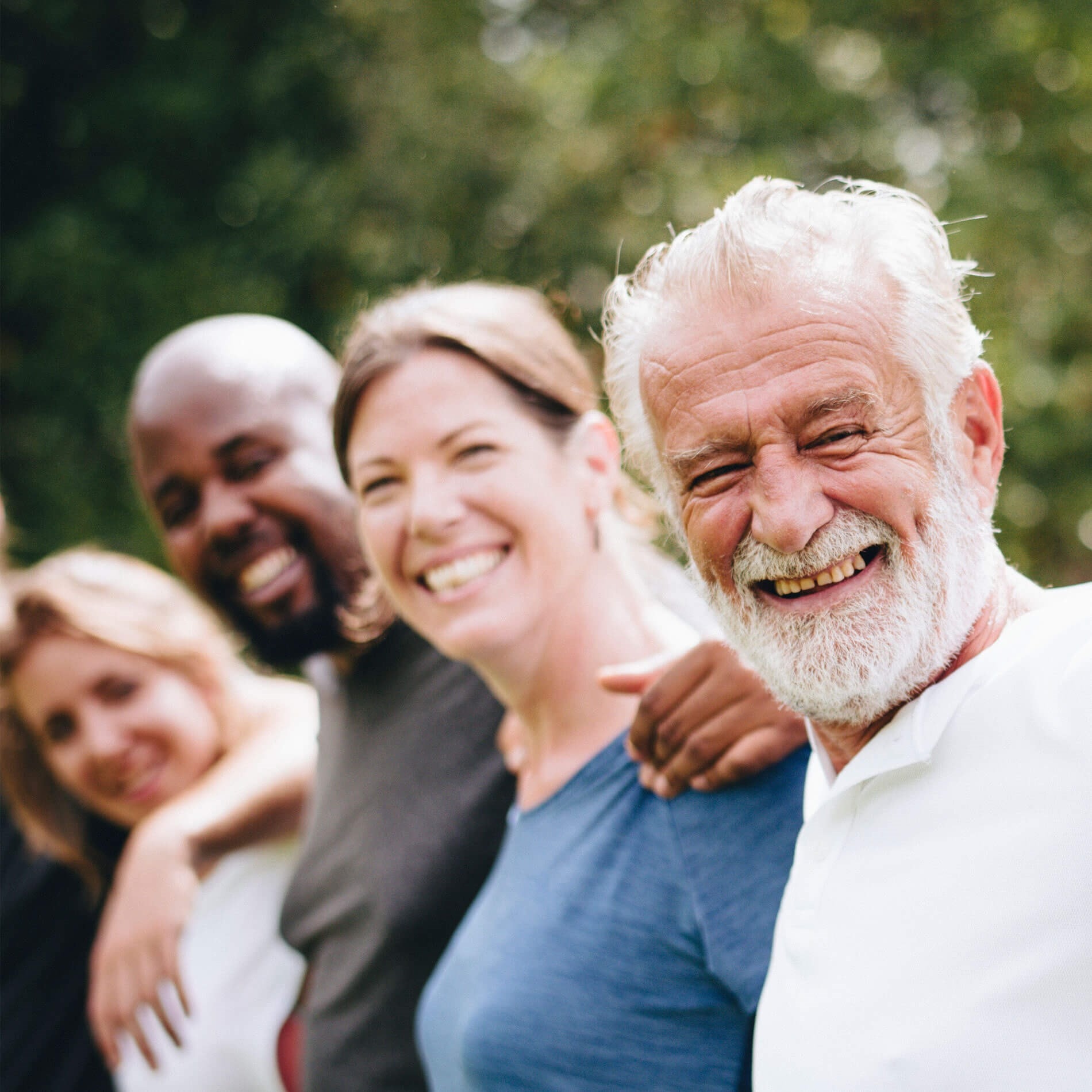 A diverse group of people smiling outdoors, standing closely together with arms around each other, with greenery in the background.
