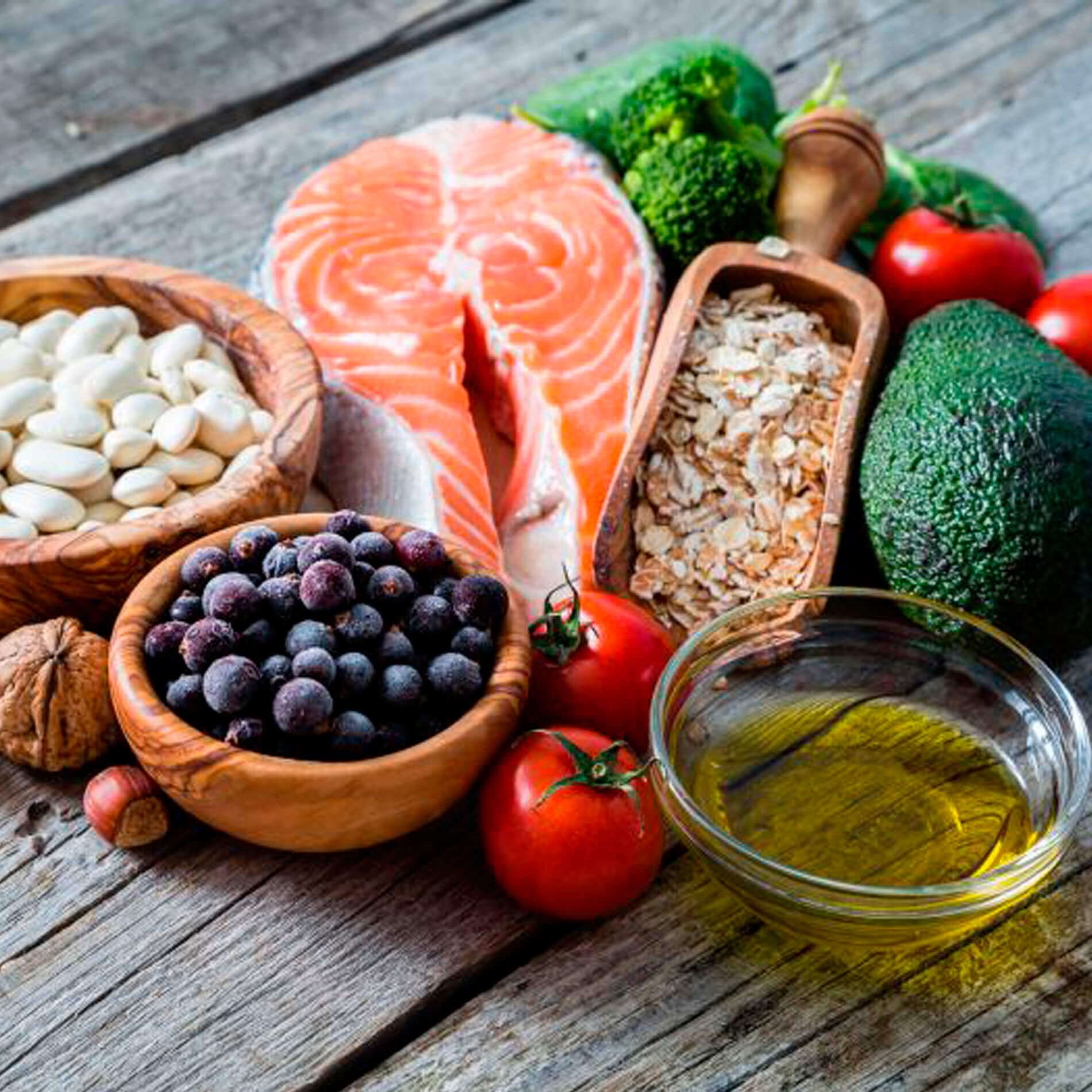 Assorted healthy foods on a wooden table, including salmon, beans, oats, blueberries, avocado, tomatoes, broccoli, walnuts, and olive oil.