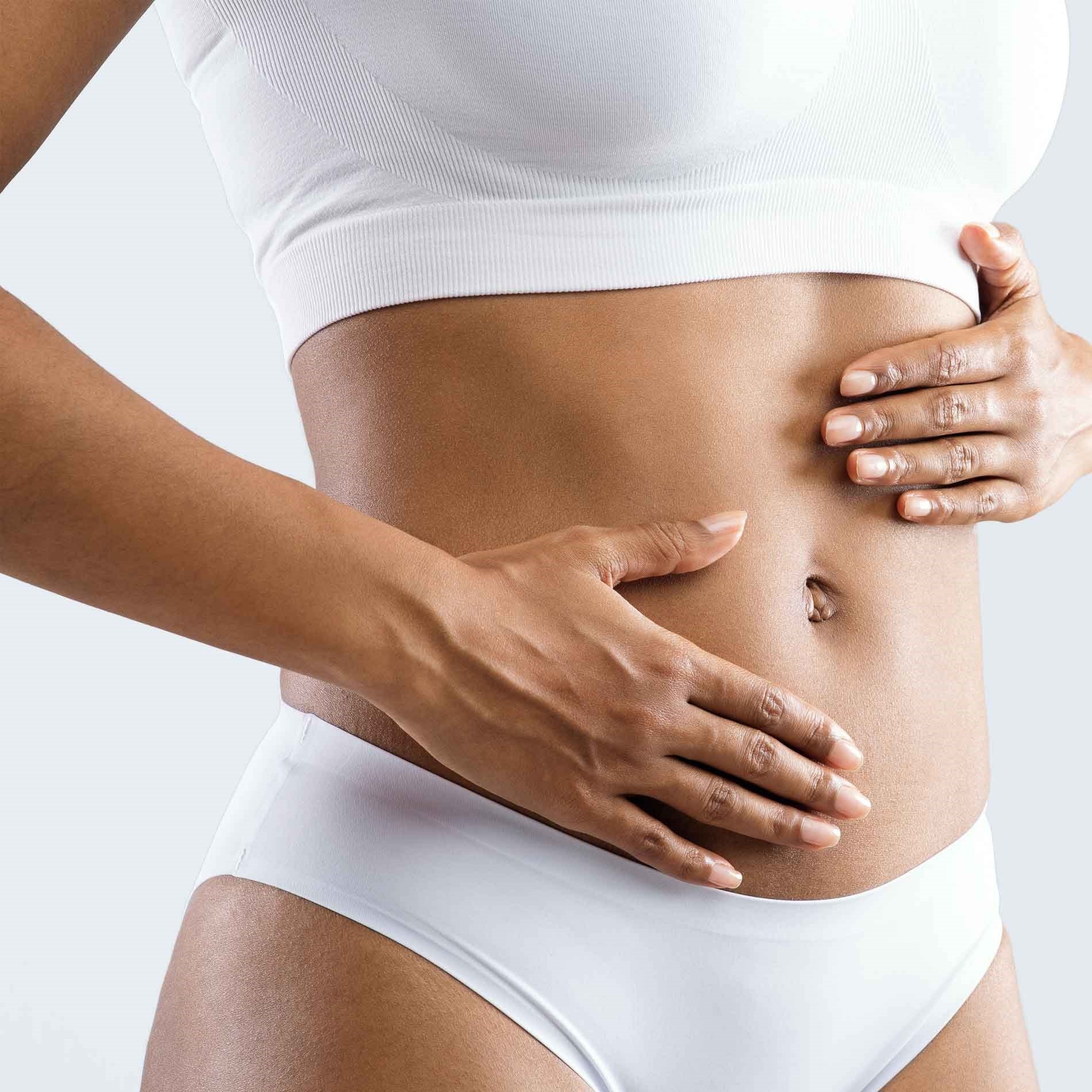 Person in a white sports bra and underwear gently touching their abdomen against a light gray background.