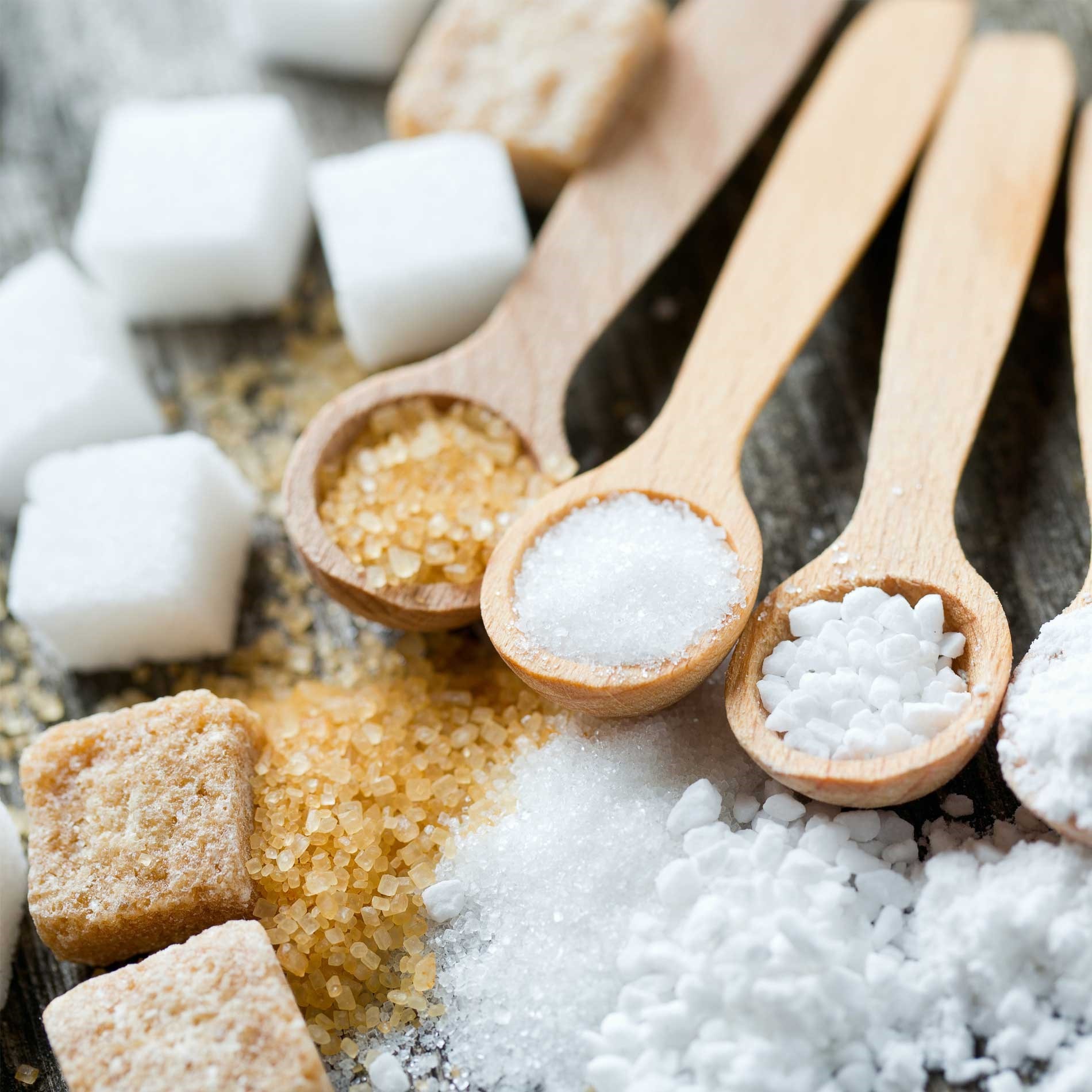 Wooden spoons with various types of sugar, including white, brown, and powdered, surrounded by sugar cubes on a wooden surface.