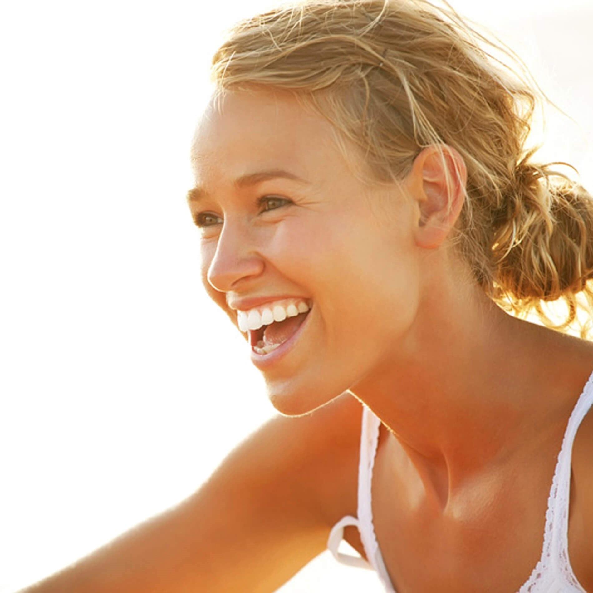 Smiling woman with blonde hair in a ponytail, wearing a white tank top, enjoying a sunny day outdoors.
