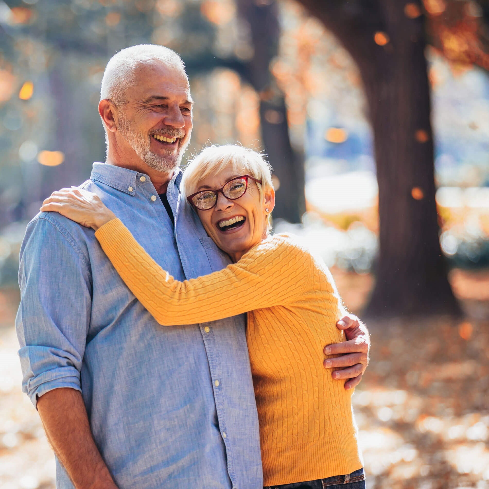 Elderly couple embracing and smiling in a sunlit park with autumn leaves falling around them.