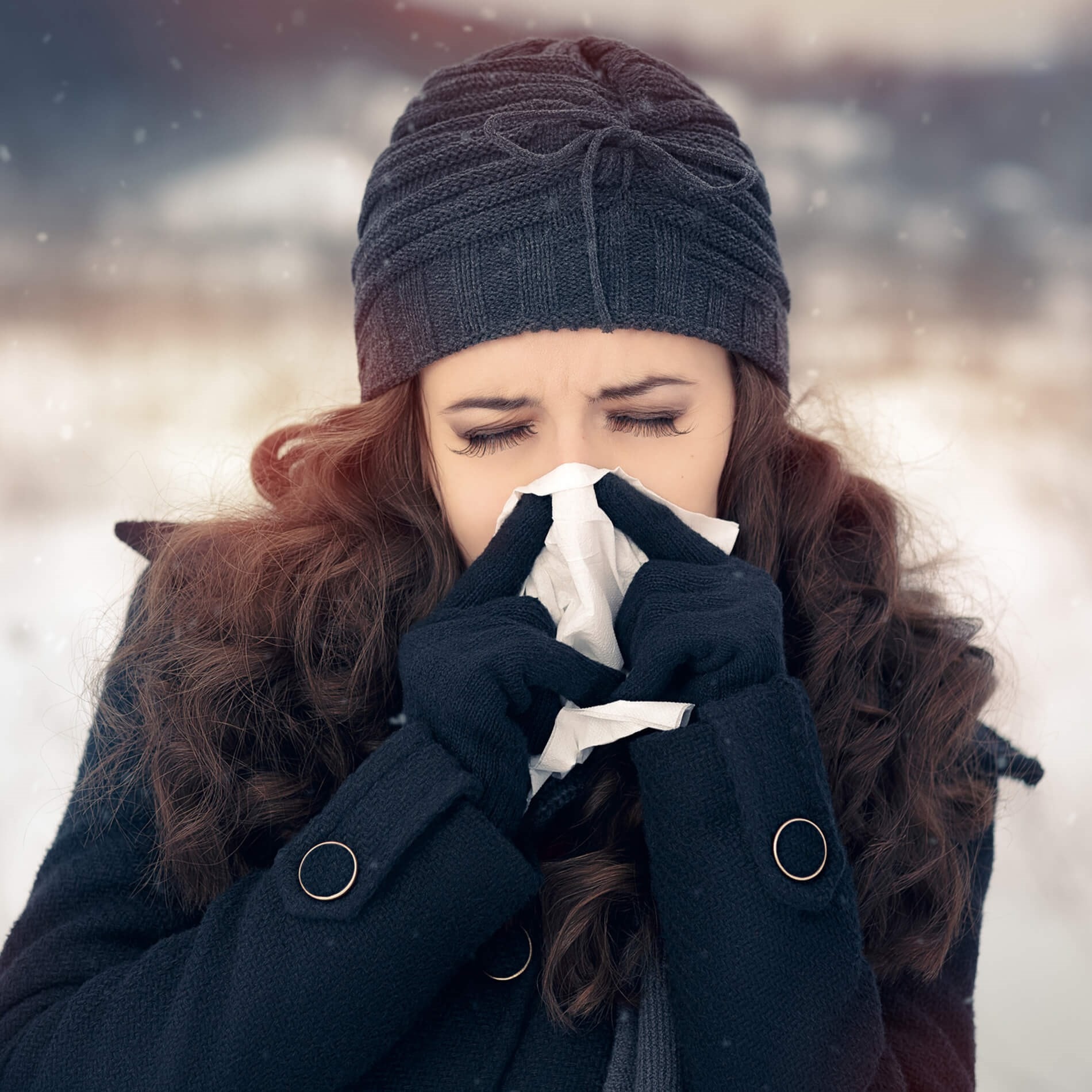 Woman in winter clothing sneezing into a tissue outdoors, with snow gently falling around her.