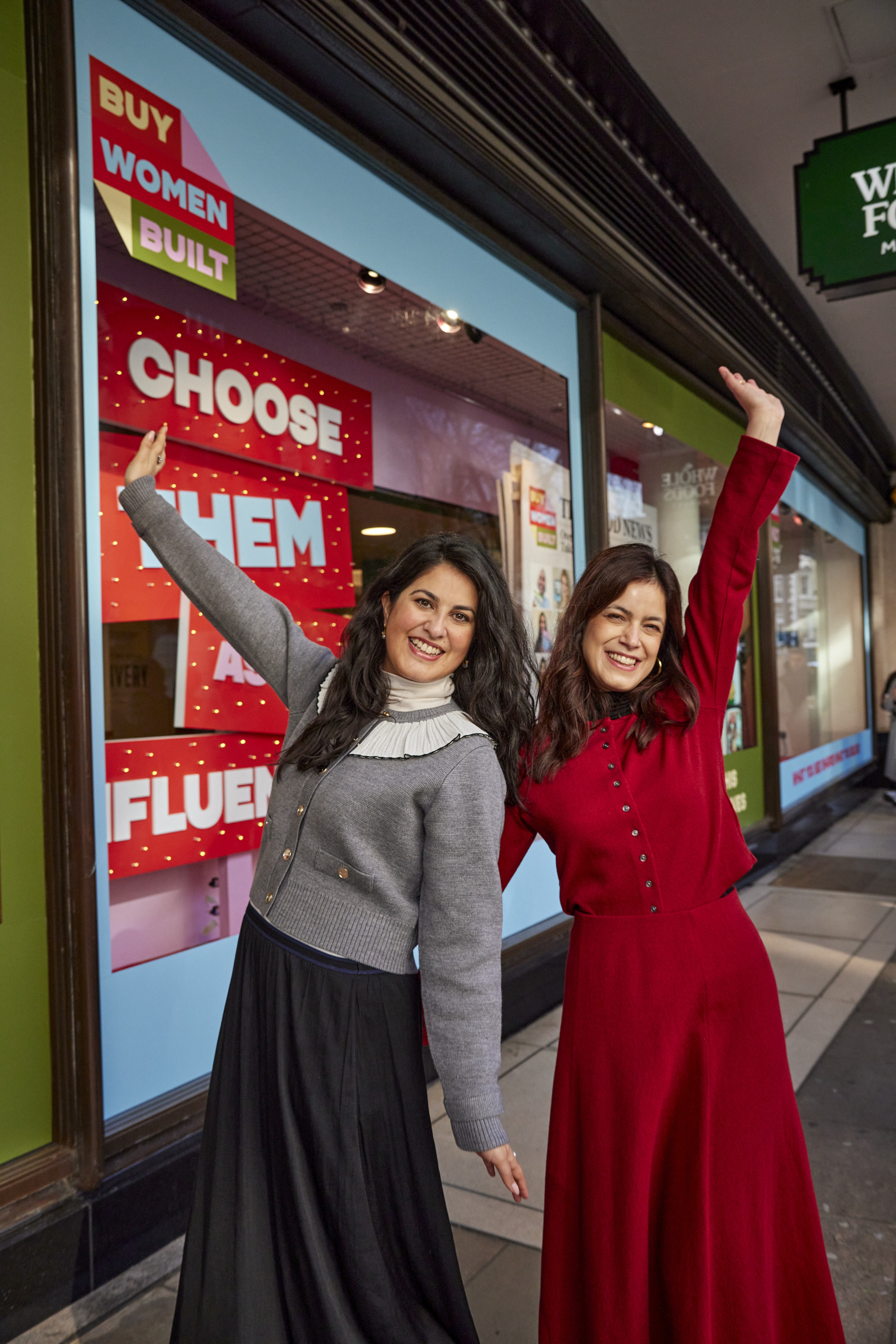 Two smiling women stand with arms raised in front of a colorful storefront with signs promoting women-built brands.