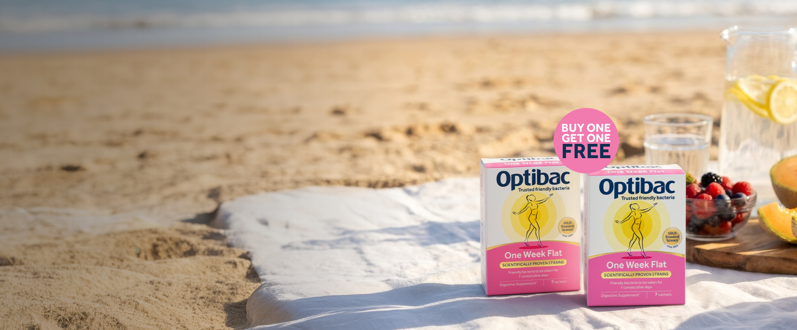 Optibac "One Week Flat" probiotic boxes on a beach picnic setup with fruit, water, and sand in the background. "Buy One Get One Free" label visible.