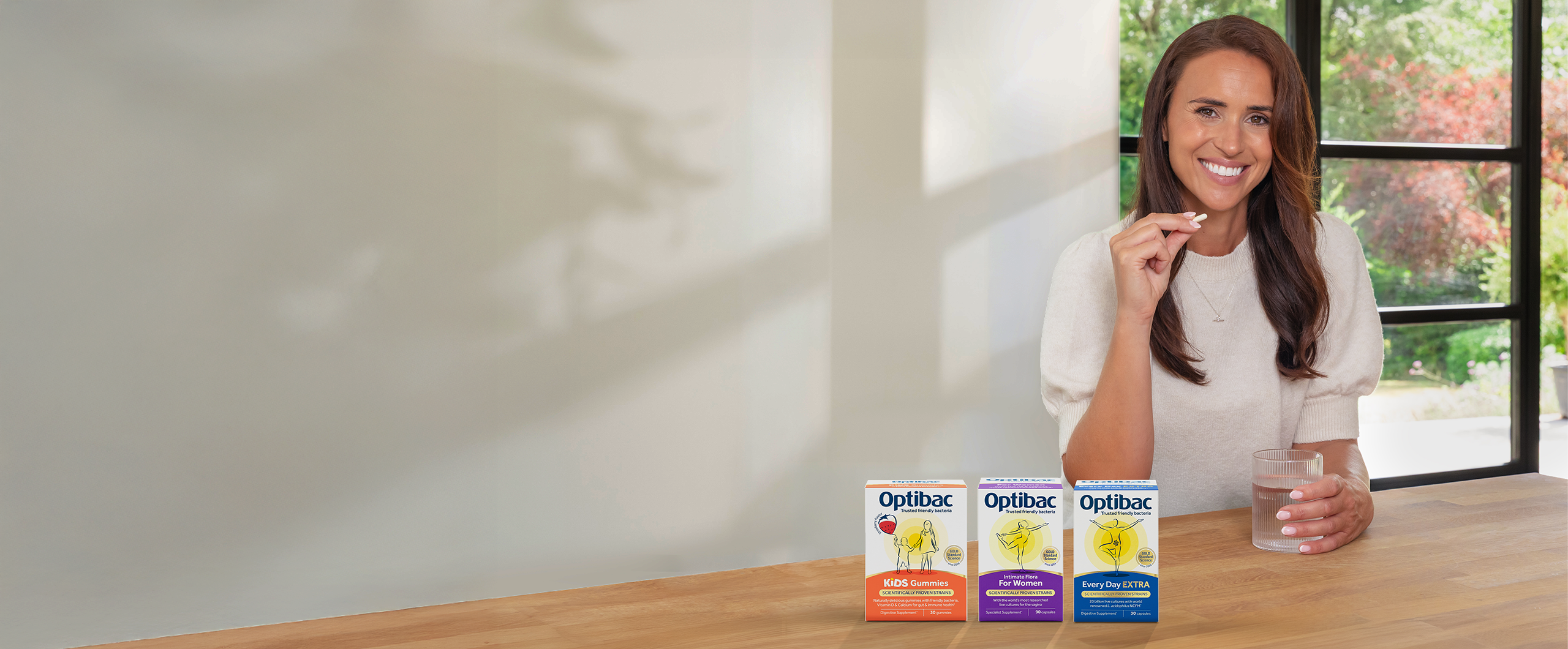 A woman smiling, holding a probiotic and a glass of water, with three boxes of Optibac probiotics on the table in front of her.