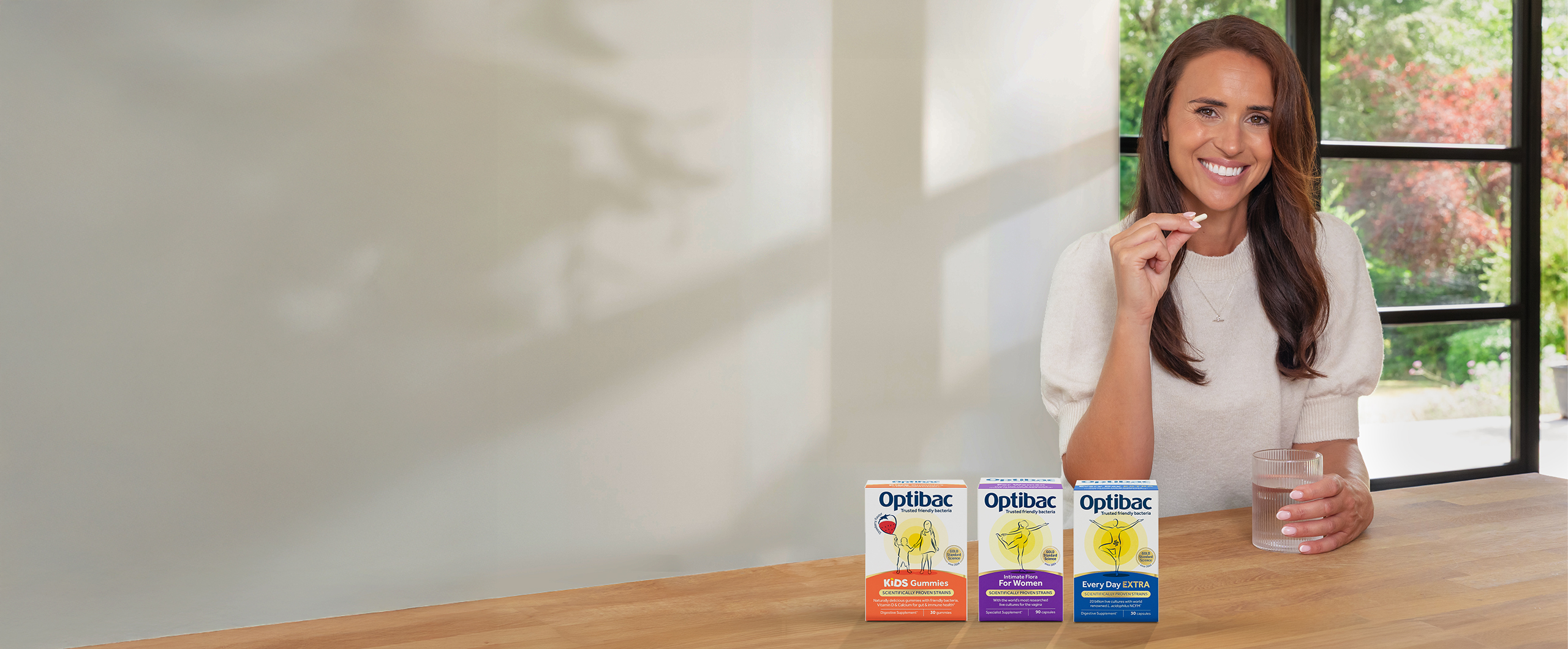 Woman smiling, sitting at a table with three Optibac probiotic boxes, holding a capsule and a glass of water, with a garden view in the background.