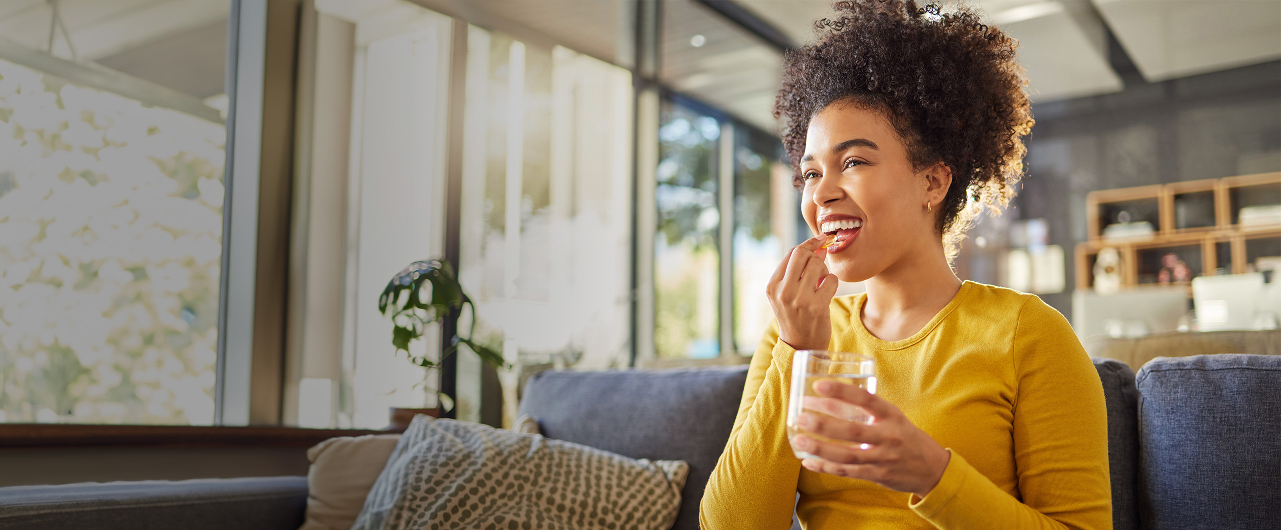 Woman in a yellow sweater sits on a couch, smiling while eating a snack and holding a glass of water in a sunlit living room.