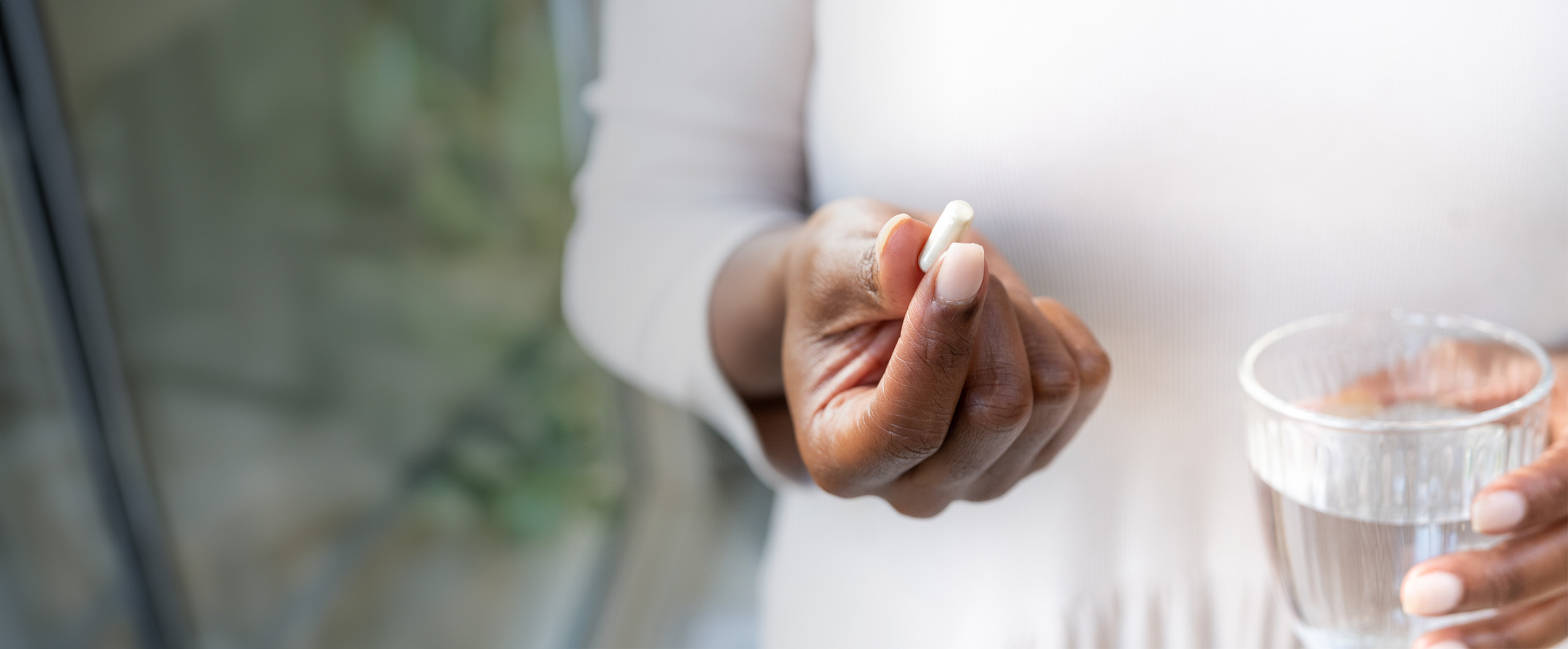 Person holding a pill in one hand and a glass of water in the other, standing near a window.