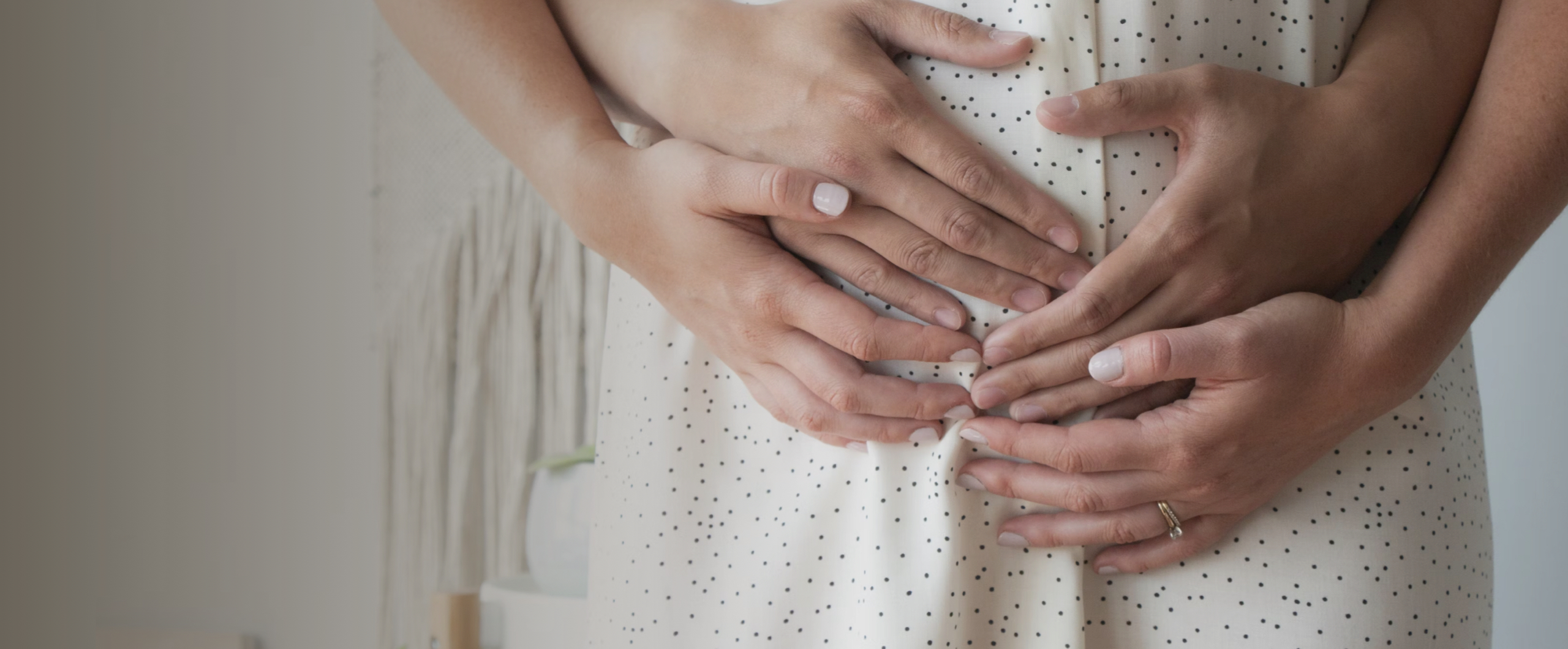 Two pairs of hands gently rest on a pregnant belly, covered by a white polka-dotted dress, symbolising love and anticipation.