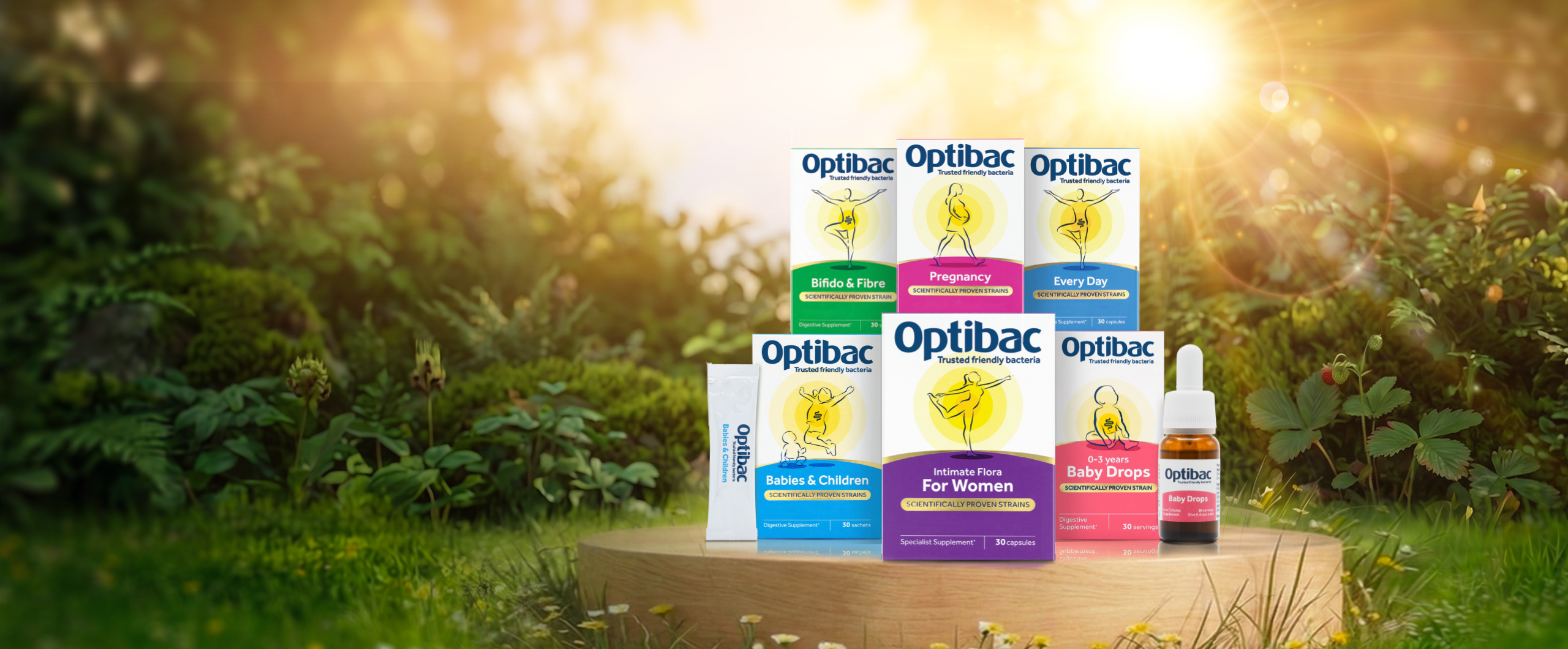 Optibac probiotic products displayed on a wooden surface in a sunny, lush garden setting.