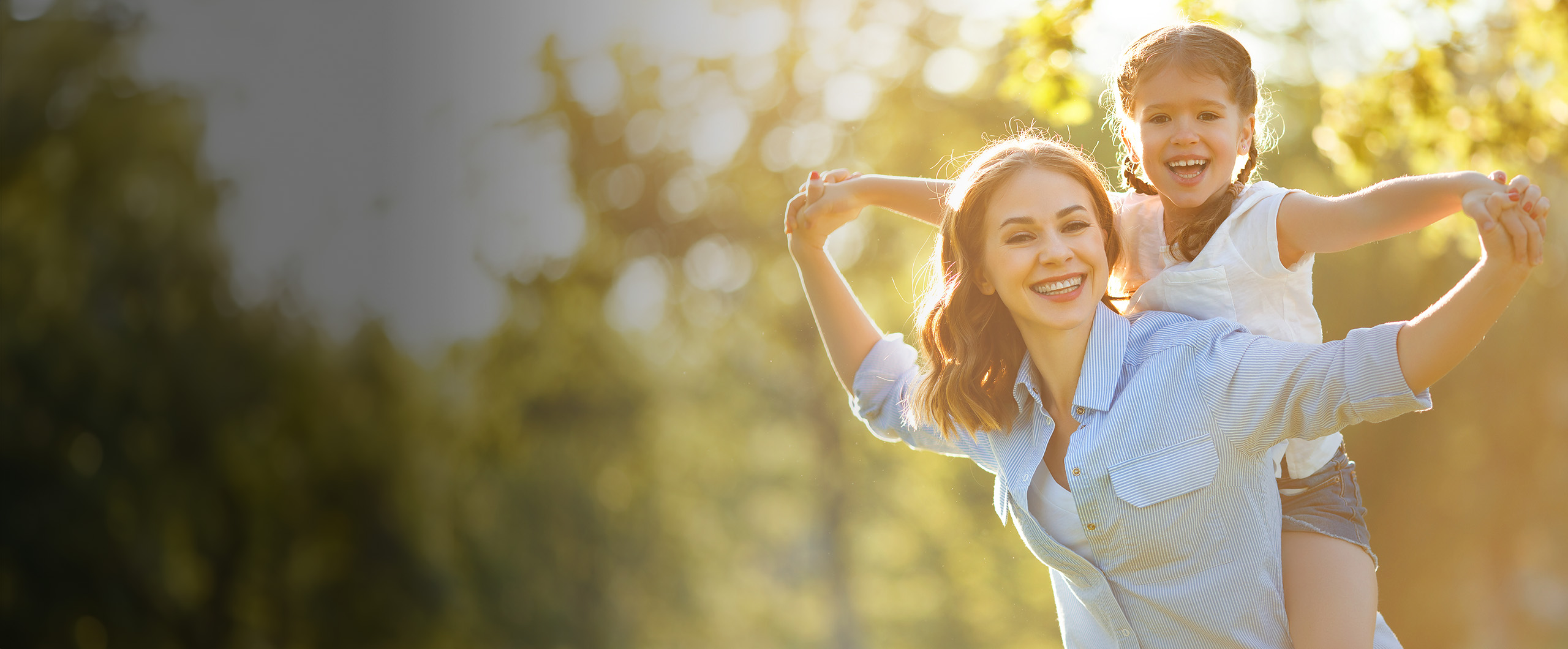 A smiling woman gives a piggyback ride to a joyful girl in a sunny park, surrounded by green trees and a warm, golden glow.