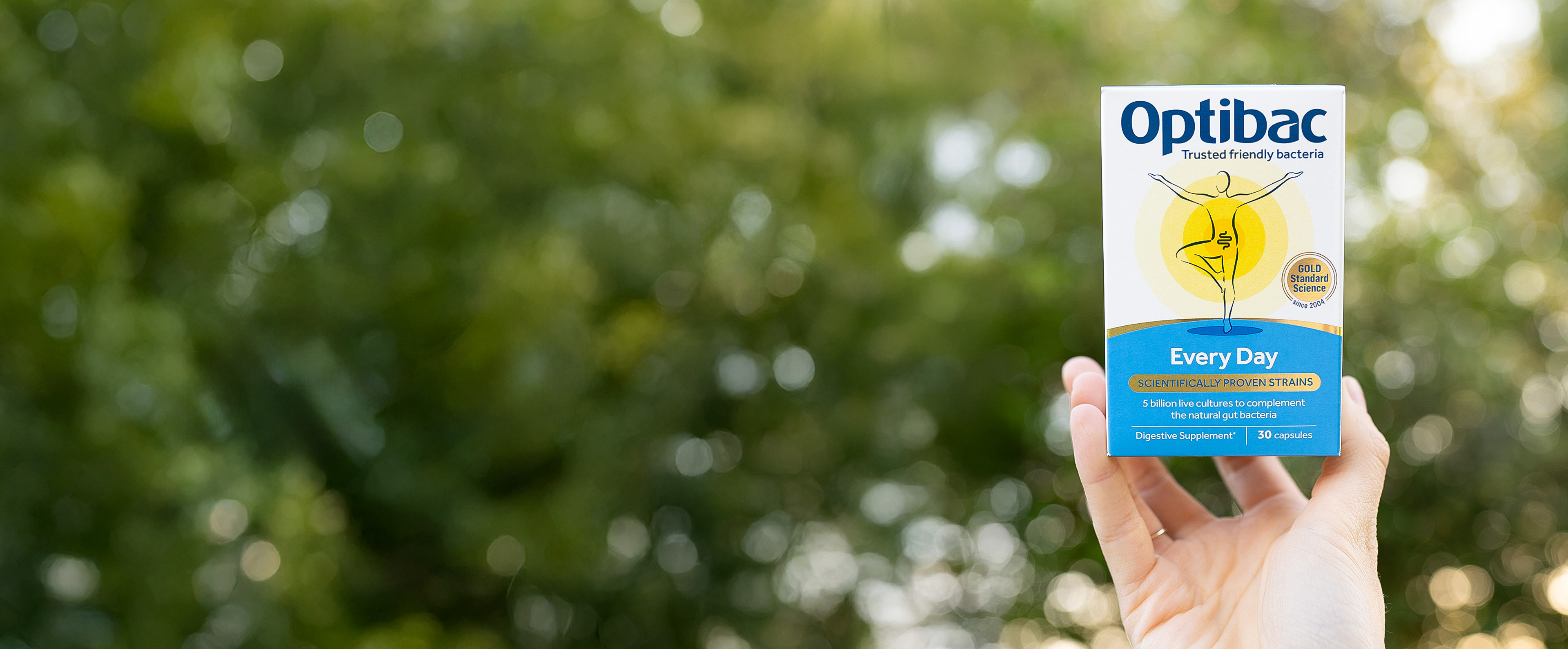 Hand holding a box of Optibac Every Day probiotic supplement against a blurred outdoor background with green foliage.
