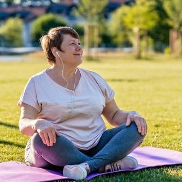 Woman sitting cross-legged on a yoga mat outdoors, wearing earphones, looking up, with trees and grass in the background.