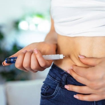 Person injecting medication into their abdomen, wearing a white top and jeans, with a blurred background.