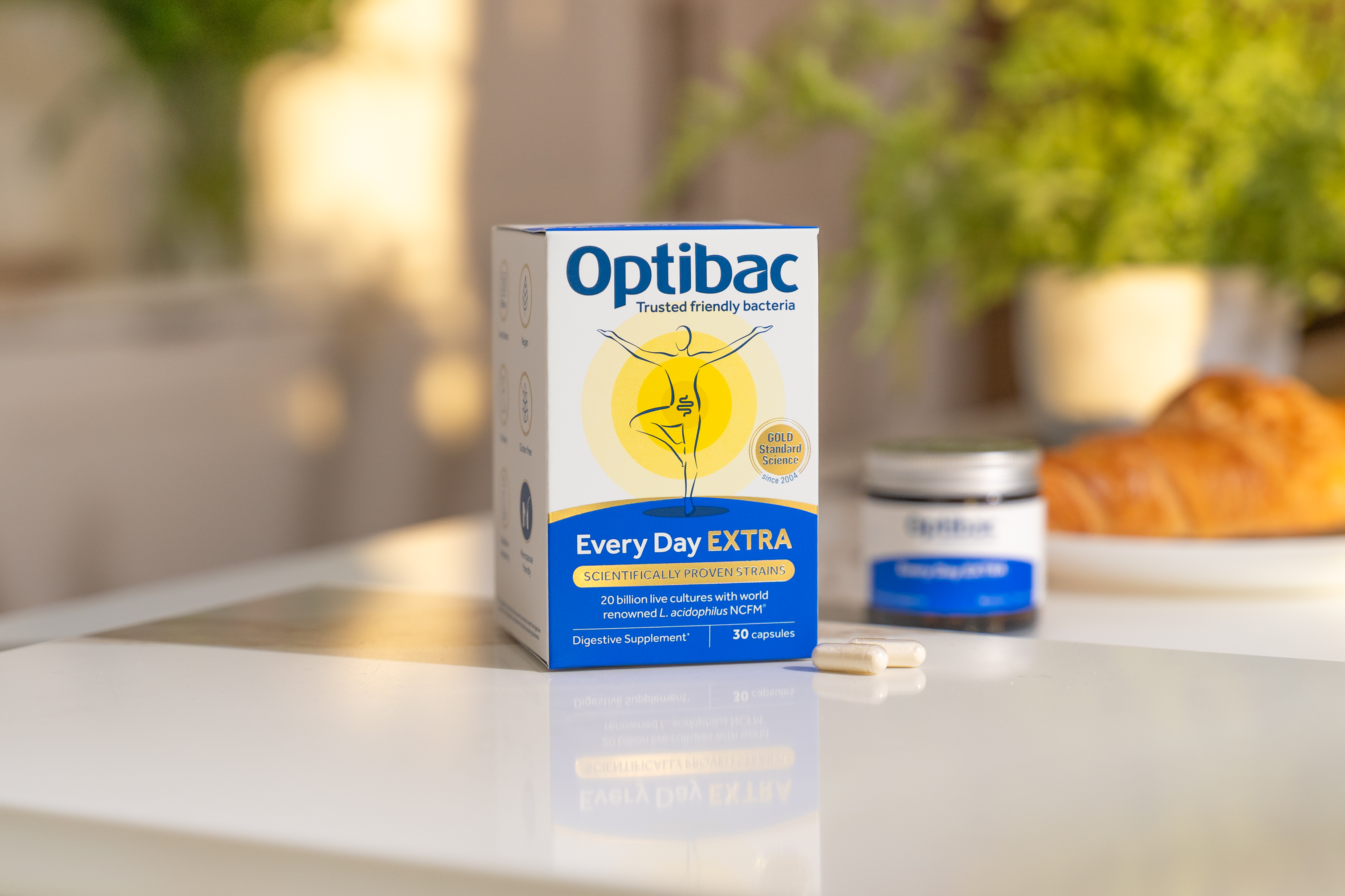 Optibac Every Day EXTRA probiotic box on table with capsules, green plant, and croissant blurred in the background.