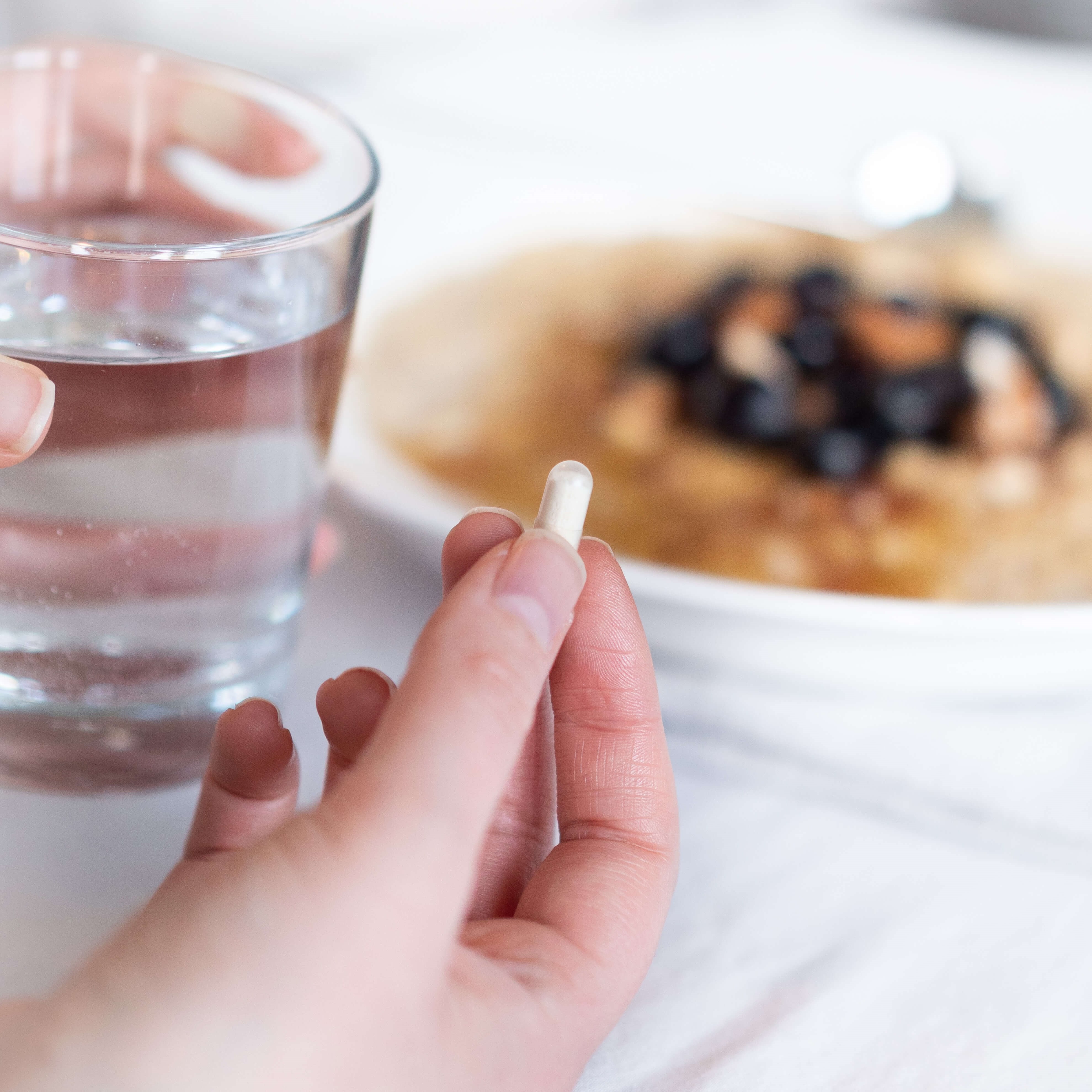 Hand holding a white capsule with a glass of water, plate of food blurred in the background on a white table.