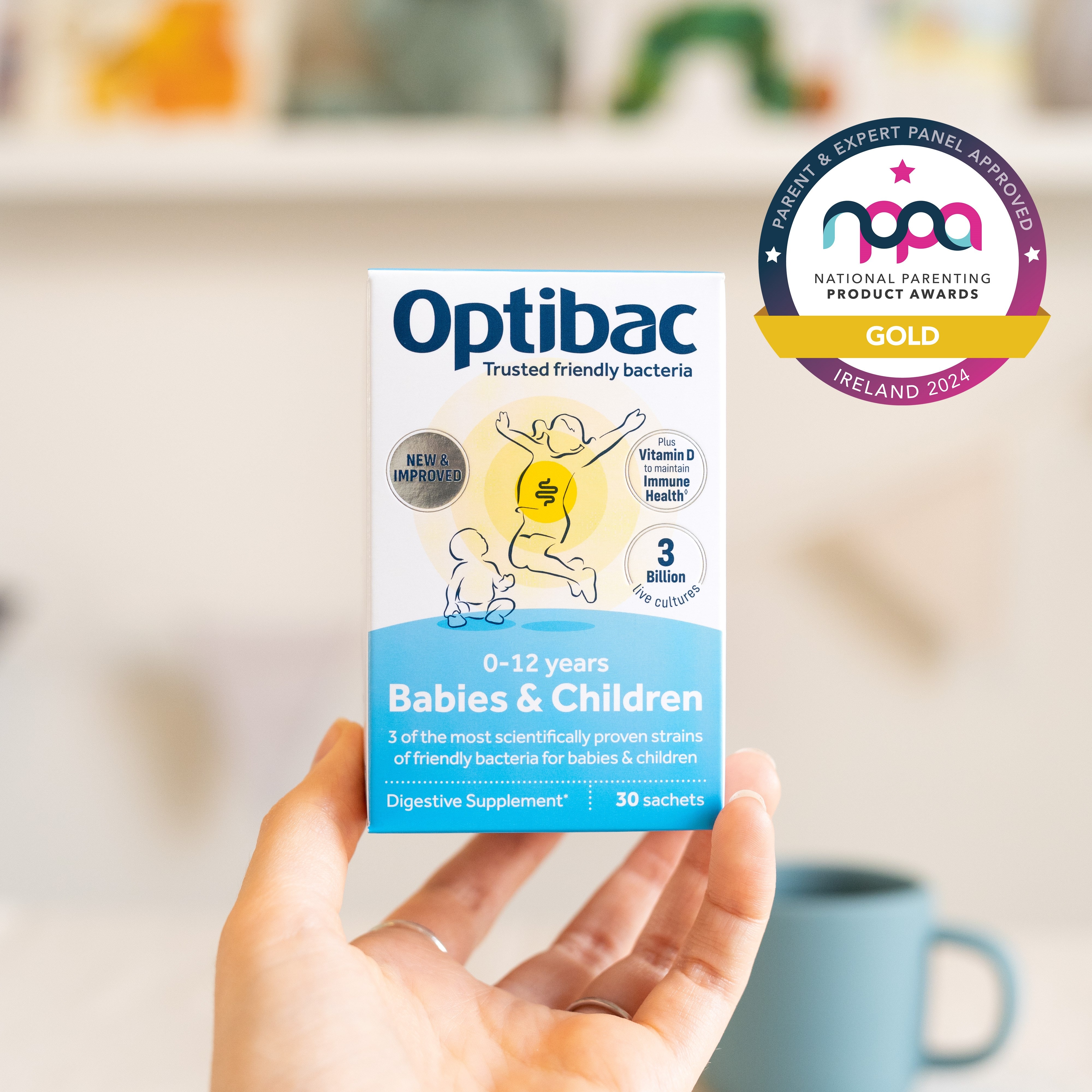 Hand holding a box of Optibac Babies & Children digestive supplement. The box highlights vitamins D and B6, with award badge visible.