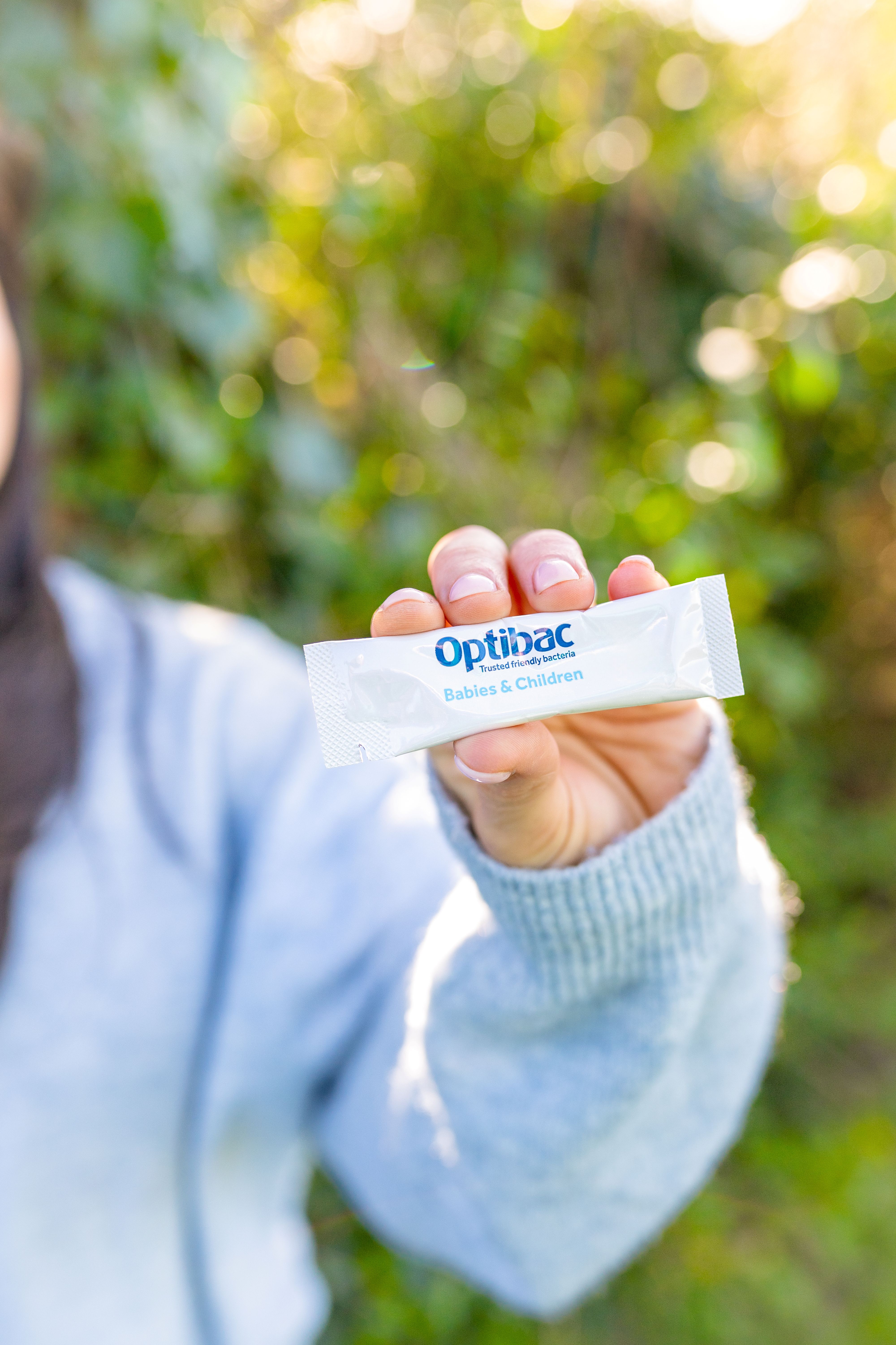 A person holds up a sachet labeled "Optibac Babies & Children" outdoors with a blurred green background.