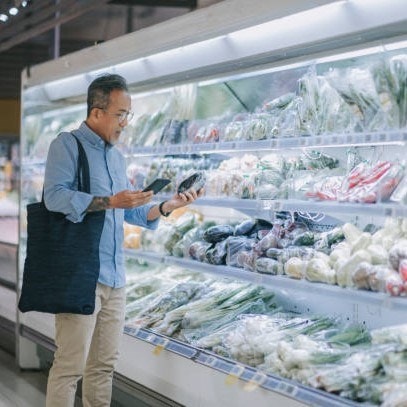 Man shopping for vegetables in a grocery store, holding produce and smartphone, with a reusable bag over his shoulder.