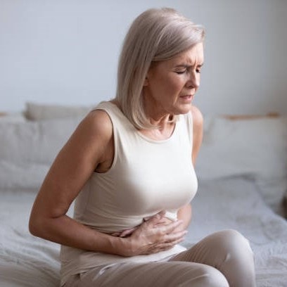 Woman with light hair sitting on a bed, wearing a beige outfit, holding her stomach with a pained expression.