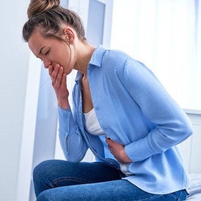 Woman in a light blue shirt and jeans, sitting and holding her stomach with one hand, covering her mouth with the other, appearing unwell.