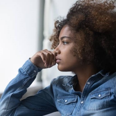 Person with curly hair in a denim shirt, sitting thoughtfully with their chin resting on their hand, gazing to the side.
