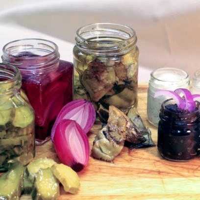 Jars of pickled vegetables, including artichokes and onions, on a wooden board with sliced red onion and a small jar of dark olives.